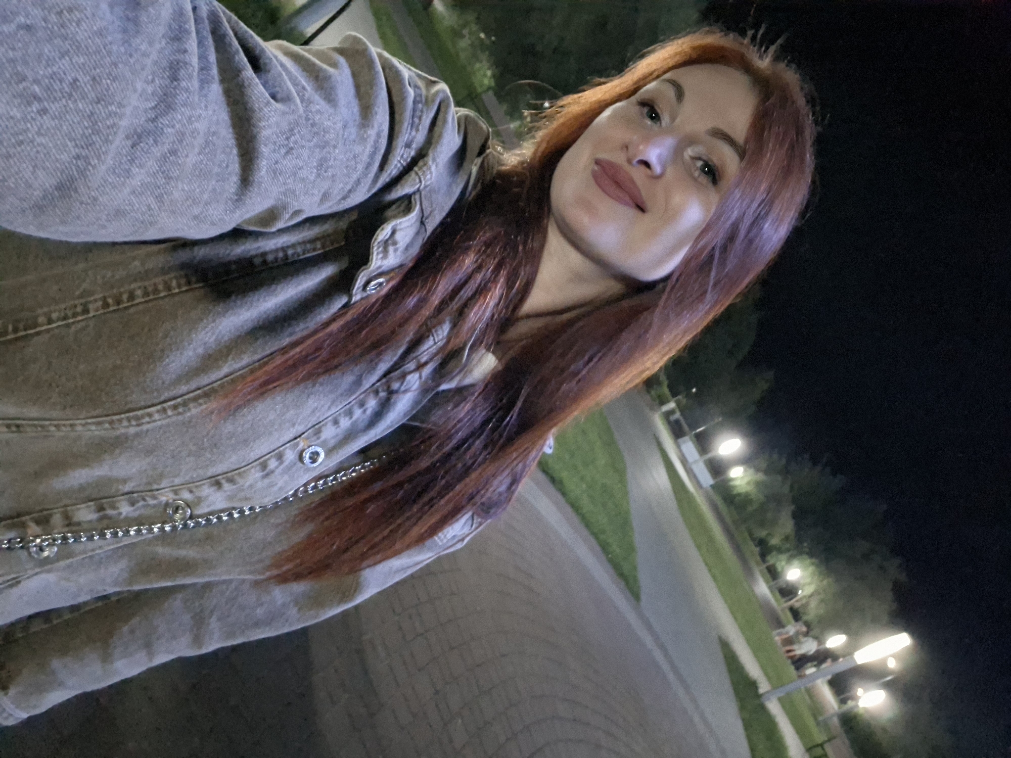 The image shows a young woman with long, reddish-brown hair and fair skin posing for a selfie at night in a park. She is wearing a grey denim jacket with silver buttons and a silver chain strap visible over her chest. She has makeup on and is smiling faintly. The background is dimly lit by streetlights and appears to be a park pathway with trees and grass. There are a few people in the background, some of whom are near a streetlight. The ground is paved with interlocking stones in a curved pattern. The overall setting is outdoors in an urban park environment at night. Based on the provided context, this scene is located in the city of Myadzyel, Belarus.