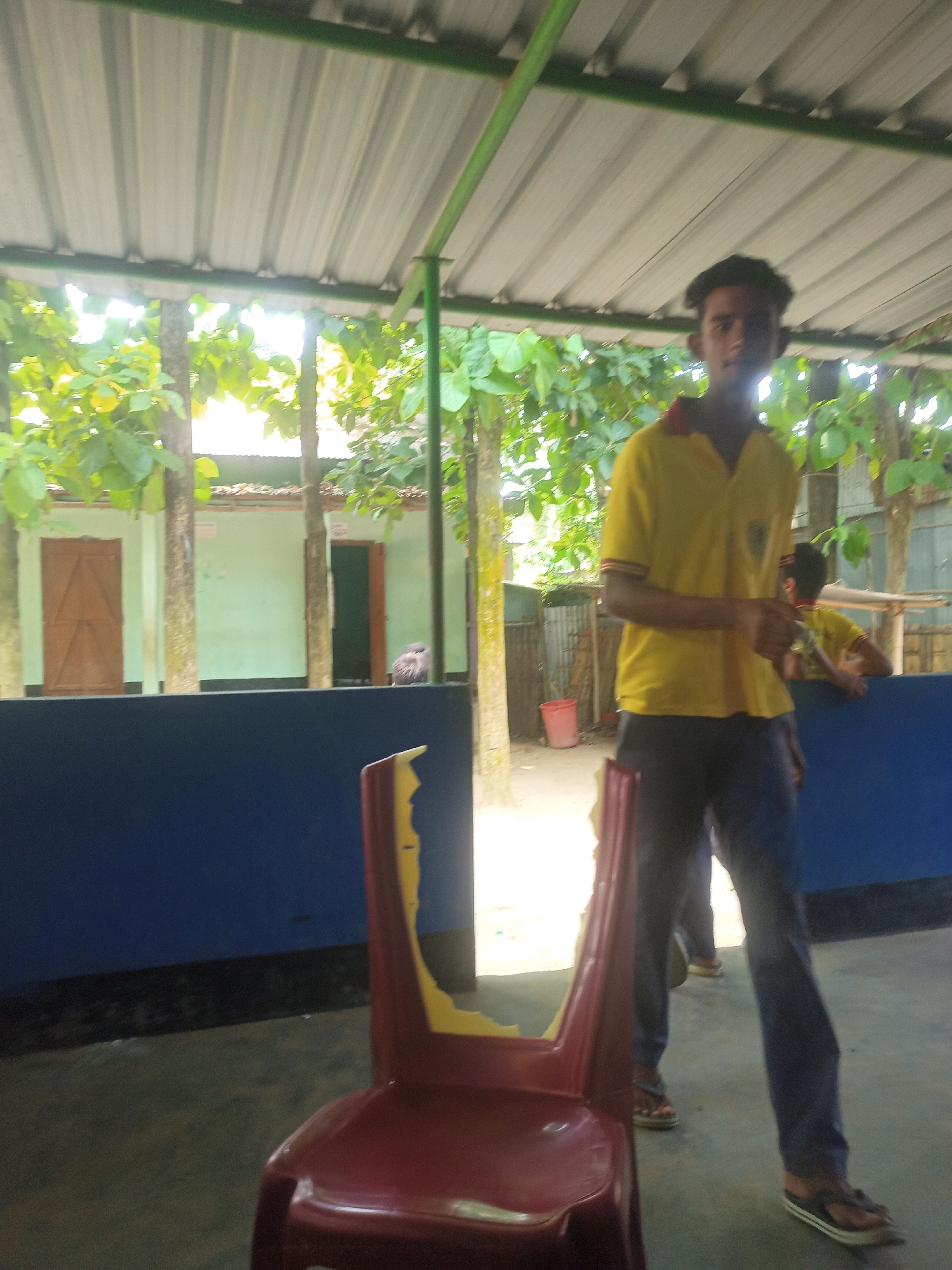 A young man in a yellow polo shirt and dark pants stands in front of a red chair. He is facing the camera and appears to be looking at it. In the background, there is a building with light green walls and brown doors. There are trees with green leaves to the left and right of the building. The scene appears to be outdoors, possibly in a schoolyard or courtyard. The image is taken from a low angle, and the man is slightly out of focus. The specific location in the city of Hatsinigmari, India is not determinable from the image.