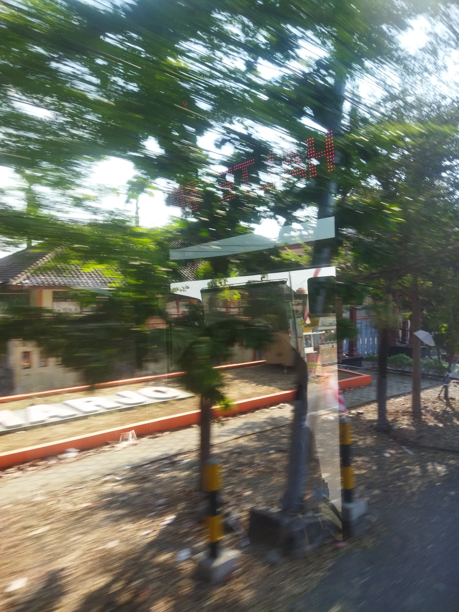The image depicts a blurry outdoor scene with motion blur, suggesting movement. In the foreground, a paved surface with scattered debris is visible. A striped pole, black and yellow, stands on the right. To the left, large white letters spelling "ARUJO" are partially obscured by motion blur, set against an orange border. Behind this, there are buildings with tiled roofs, surrounded by lush green trees. A metallic structure with reflective surfaces, possibly a bus or vehicle, is visible in the middle ground, showing reflections of the surroundings. Red LED lights are also discernible in the upper right portion of the image, arranged in a pattern that suggests a sign. The overall impression is of passing by a suburban or semi-urban area in daylight, with significant motion blur indicating a fast-moving perspective. The presence of the name "ARUJO" suggests this location might be within or near a place associated with that name, potentially a landmark, street, or business.