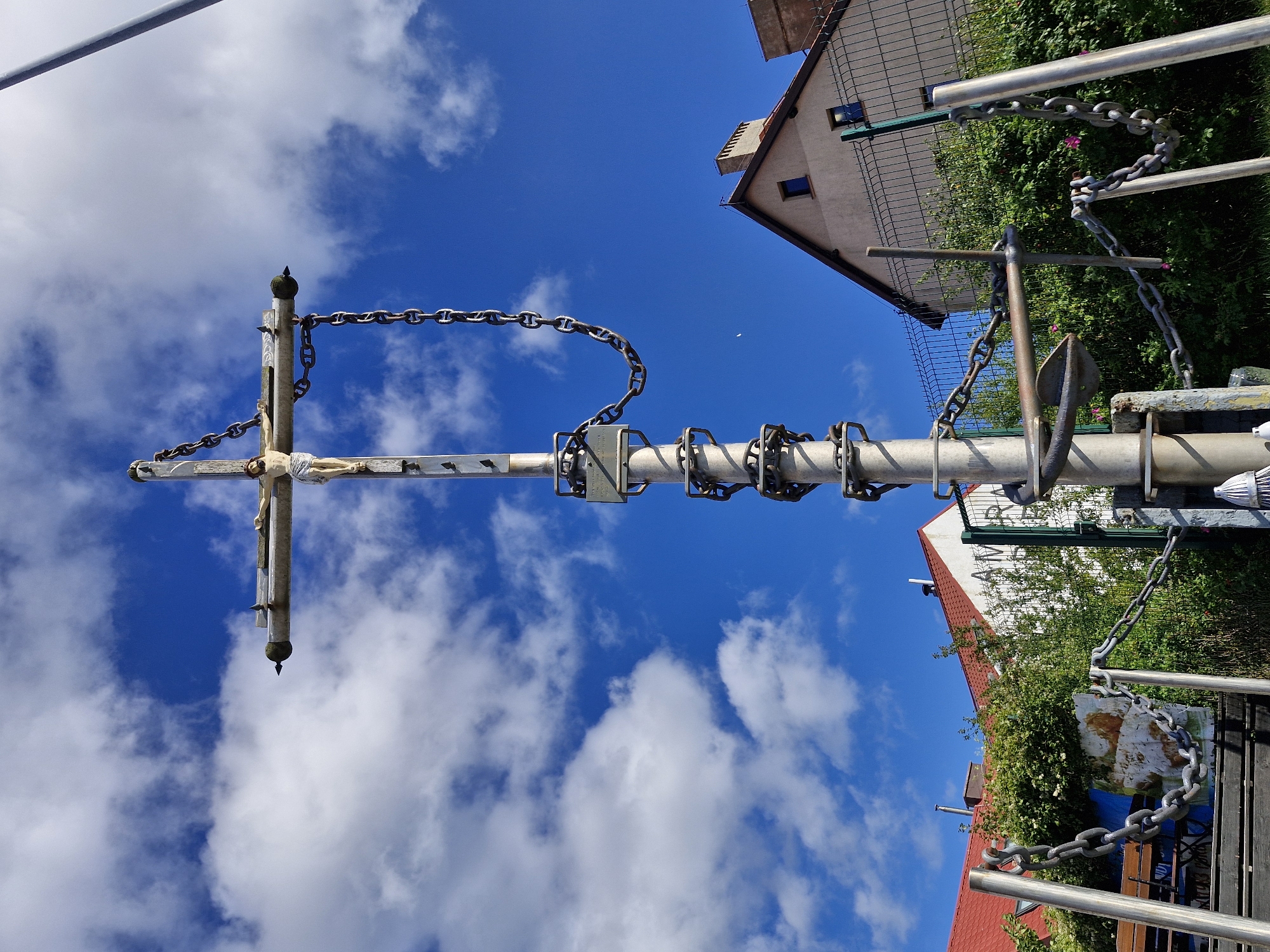 A low-angle, full shot captures a tall, metallic cross topped with a figure of Jesus Christ, against a bright blue sky dotted with white clouds. The cross, made of weathered material, features thick metal chains wrapped around its vertical pole and extending downwards to posts in the foreground. A small plaque is visible on the pole. The cross is situated in what appears to be a public space, with a fence, lush green bushes, and the roofs of residential buildings visible in the background. The buildings have red-tiled roofs and light-colored walls, one with a visible chimney. The overall impression is of a monument or religious marker in a town setting, possibly on a sunny day. The scene is located in the city of Hel, Poland.