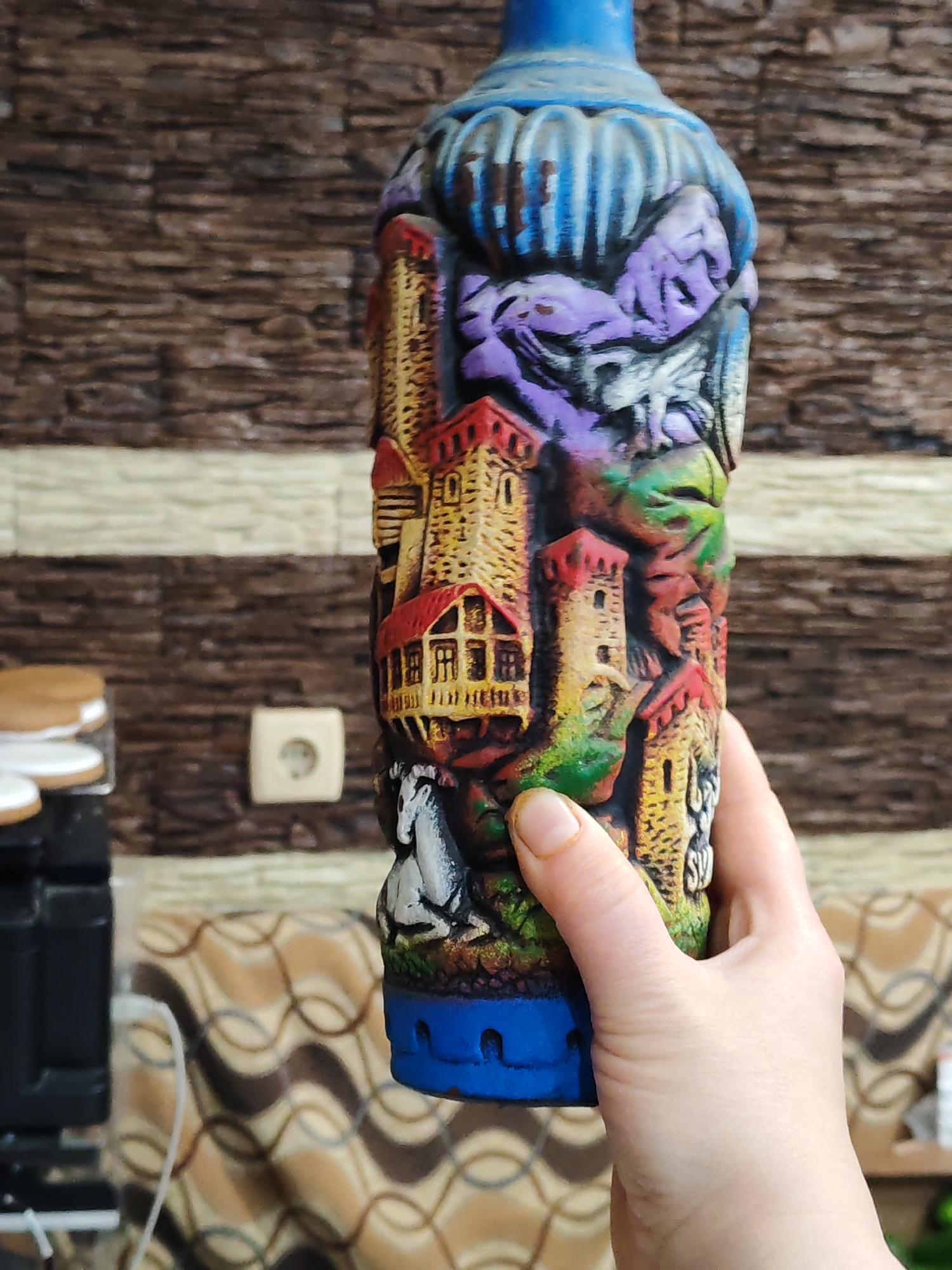 A hand is holding a tall, ornate ceramic bottle. The bottle features a detailed, raised design of castles, horses, and what appears to be mythical creatures. The bottle is painted in multiple colors, including blue, purple, green, red, and yellow. The background is blurry, but a textured wall and furniture can be discerned. The location and city are Unknown.