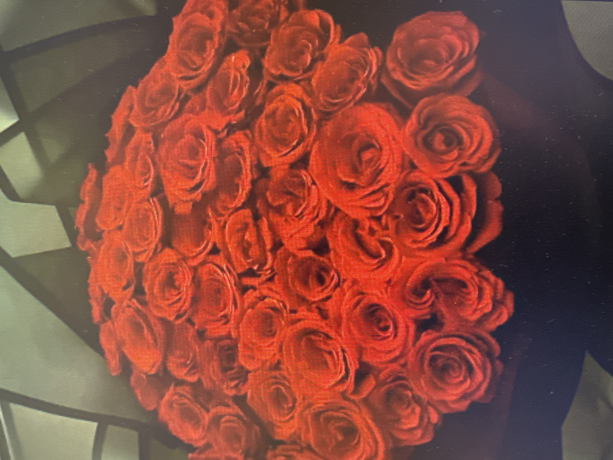 The image shows a close-up view of a large bouquet of red roses. The roses are tightly packed together, with many layers of petals visible. The lighting is somewhat dim, creating shadows that accentuate the depth and texture of the blooms. The background is dark and out of focus, making the vibrant red roses the central subject. There are no people or any identifiable landmarks in the image.