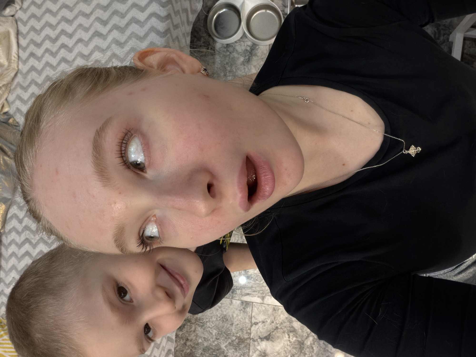 The image shows two fair-skinned individuals in an indoor setting. One is a young adult woman with blonde hair wearing a black shirt and a silver necklace with a pendant. The other is a child with light brown hair, also wearing a dark shirt. They appear to be taking a selfie or posing for a photograph together.
The background includes a gray and white patterned surface (possibly a bedspread or blanket), a gray tile floor, and two small, round metal bowls or dishes.
The image's location within Rossiyskiy, Russia, cannot be determined with the given information.