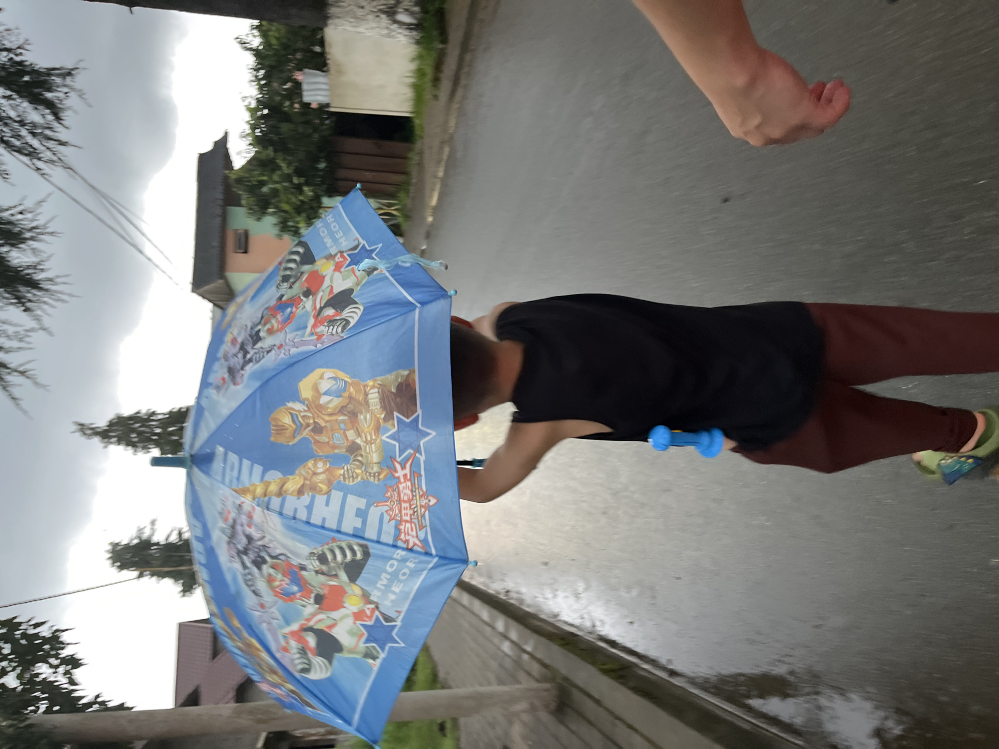 A young person, viewed from behind, holds a blue umbrella with animated characters on it. They are wearing a black sleeveless shirt and maroon pants. They appear to be walking on a wet paved surface, suggesting it is raining. A second person's arm is visible in the lower right of the frame, reaching out towards the viewer. The background shows buildings, trees, and a cloudy sky, consistent with a residential area in Ureki, Georgia.