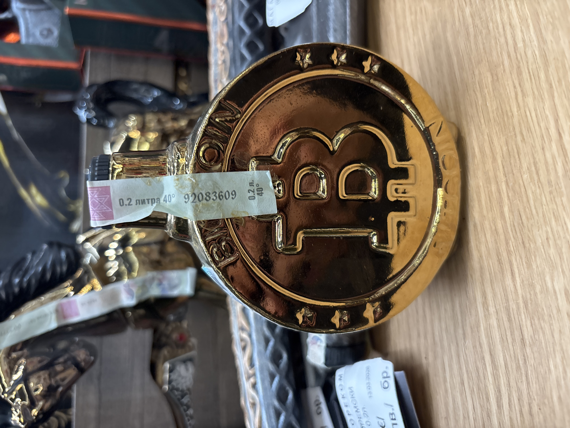Here is a description of the image:
The image shows a gold-colored bottle shaped like a Bitcoin, lying on a wooden surface. A label is attached to the neck of the bottle, with the text "0.2 литра 40° 92083609". The bottle appears to be a souvenir or novelty item, likely containing liquor given the "40°" marking. The text "BITCOIN" is inscribed around the bottle. Other items are visible in the background. The presence of Bulgarian language text on the label and surrounding papers suggests the photo was taken in Bulgaria. The "OPEKOM СРЕМСКИ" also points towards this location, potentially within the broader Balkan region. The text "13.03.2008" is also visible on one of the labels. There is insufficient information to determine the exact location in Plovdiv, or other cities in Bulgaria.
