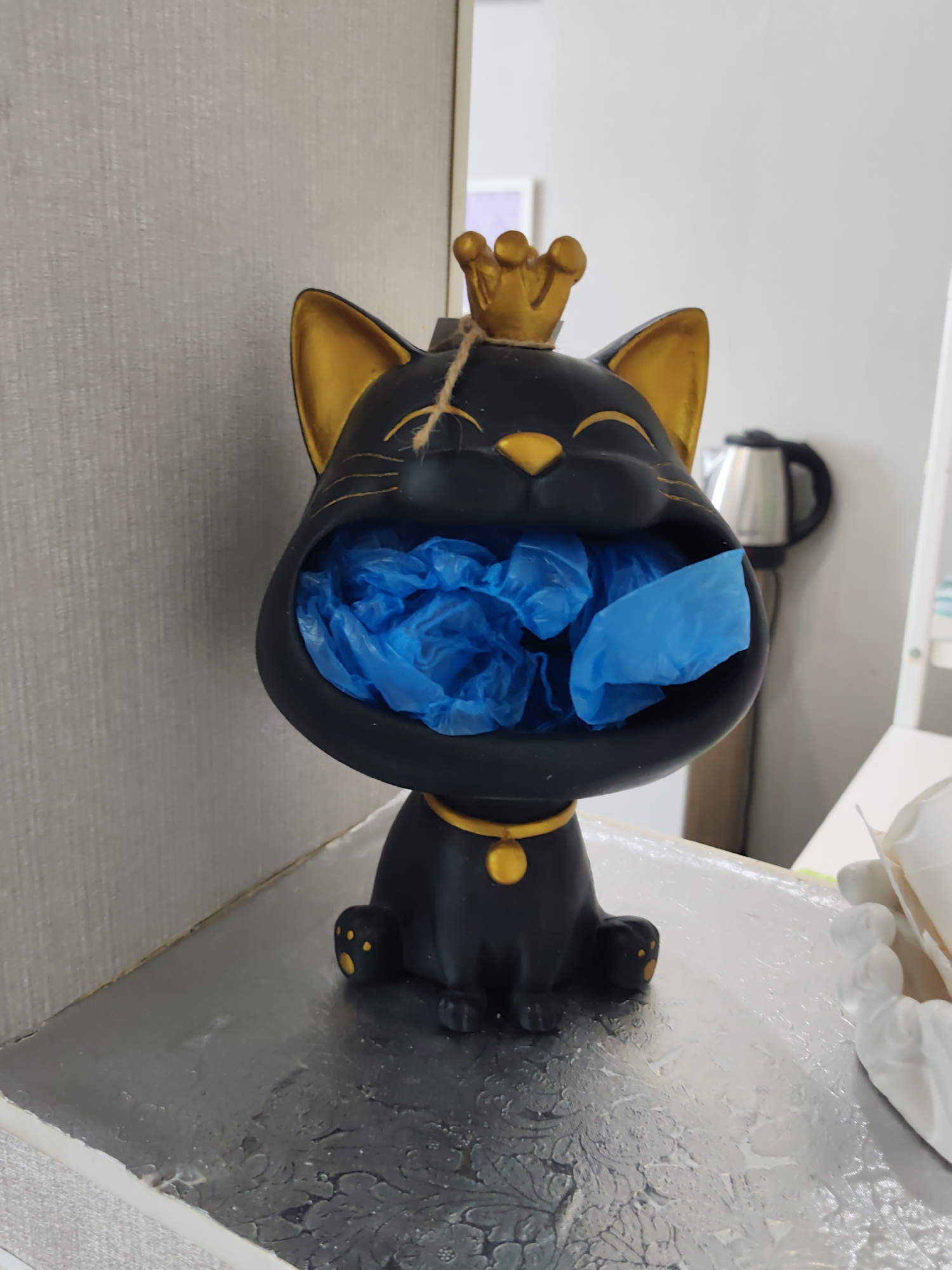A black cat figurine wearing a gold crown and collar is on a silver surface, its mouth open and filled with blue plastic bags. The figurine is positioned in front of a wall with a textured surface and to the right of a stainless steel kettle. The scene appears to be indoors, possibly in a domestic setting. The location is in the city of Dzerzhinsk, Russia.