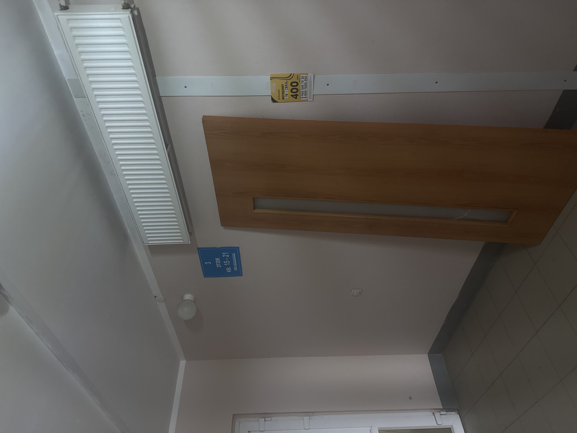 The image shows an indoor scene, likely a hallway in a residential building. A wooden door with a glass pane is visible on the right side of the frame. To the left of the door, there's a white door frame. A blue sign on the wall above the door reads "3 этаж кв. 15-21", indicating the third floor and apartments 15-21. A white radiator is mounted high on the wall above the door. A yellow flyer advertising internet and TV services for 400 units of currency is attached to a white vertical strip on the wall to the right of the door. The floor is tiled with grey square tiles. The walls are painted in a light, neutral color, and the ceiling is white. The overall environment appears to be functional and utilitarian, typical of a common area in a building. The location is identified as Kondratovo, Russia.