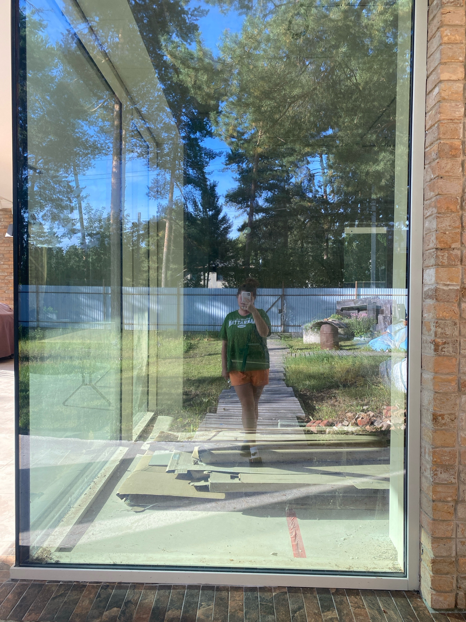 The image depicts a reflection in a large window, showing a person in a green t-shirt and orange shorts standing on a wooden pathway. The reflection reveals a backyard with a corrugated metal fence, a barrel, some plants, and piles of bricks. The overall setting appears to be a residential area, possibly a dacha or a private house. The bright sunlight suggests it is daytime. The lower part of the reflection shows building materials, such as cardboard and metal sheets, indicating ongoing construction or renovation work. The image is taken from outside the window, looking inwards at the reflection. Based on the style of the house and the vegetation, the location is consistent with a suburban or rural setting in the city of Perm, Russia.