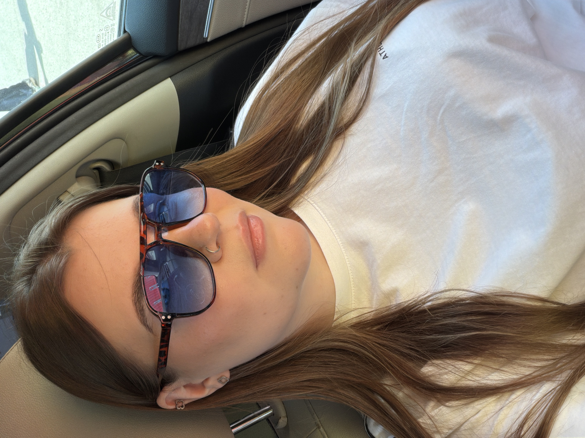 A young woman with brown hair is lying down in what appears to be a car. She is wearing sunglasses and a white T-shirt with a partial word visible on it, perhaps "ATHL". She has a nose ring and earrings. The interior of the car is visible, showing the dashboard and window frame. There is no information to determine if the image was taken in Chaikovsky, Russia.