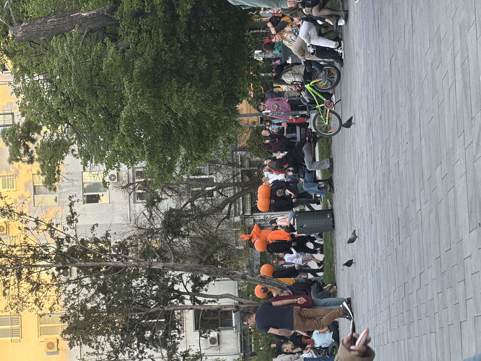 The image shows a crowd gathered on a paved area in Sofia, Bulgaria. People are sitting on benches and standing. Some individuals are holding orange balloons. There's a lime green bicycle among the crowd. Buildings are visible in the background, and trees provide shade. Pigeons are on the ground.