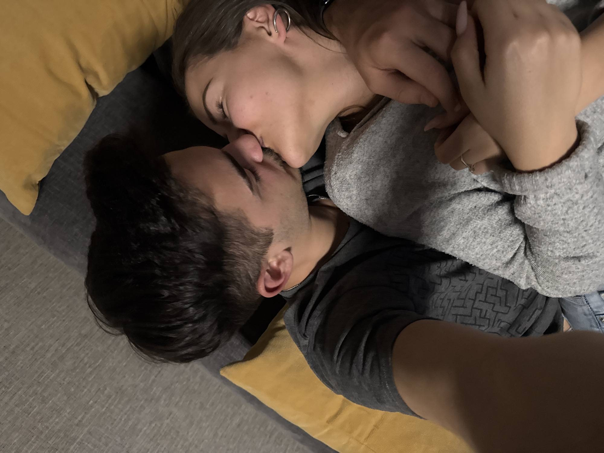 An intimate, tilted close-up shot captures a young couple lying together, engaged in a kiss. The woman is positioned above the man, her head tilted down as their lips meet. Her eyes are closed, indicating absorption in the moment, and she wears a light gray long-sleeved top, with a silver hoop earring visible in her left ear. Her hands, adorned with a ring on one finger and featuring light-colored manicured nails, are gently clasped on what appears to be the man's arm or chest area.
The man is seen from a lower angle, his face partially obscured by the woman's, with dark hair and stubble visible. He wears a dark gray t-shirt with a subtle geometric pattern and a thin chain necklace. They are reclining on a gray upholstered surface, likely a sofa or bed, with yellow pillows providing support. The lighting is soft and ambient, suggesting an indoor setting, possibly during the evening or in low-light conditions. The overall mood is one of affection and tenderness. No visible text is present in the image.