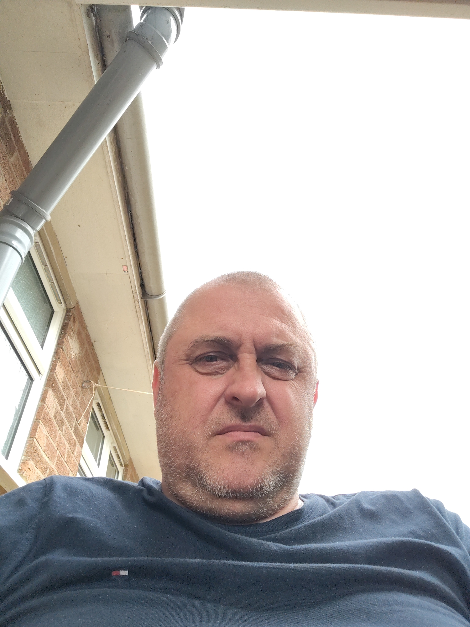 The scene is an extreme low-angle outdoor portrait of a middle-aged man, with the provided location of Huntingdonshire, United Kingdom. The man, balding with short, light grey stubble on his head and a short, grey beard, looks directly at the camera with a neutral expression. He wears a dark blue crew-neck t-shirt featuring a small rectangular red, white, and blue logo on the left chest.
Behind and above the man, the background consists of the exterior of a building. To the left, a reddish-brown brick wall is visible, along with a white-framed window. A grey plastic drainpipe descends diagonally from the upper left. Below white/cream-colored fascia boards, a grey gutter system is visible. A thin, light-colored string extends from near the window to the right, and a small, reddish-pink object (possibly a sensor or fixture) is attached to the underside of the fascia board. The upper right portion of the image is dominated by an overexposed, bright white sky.