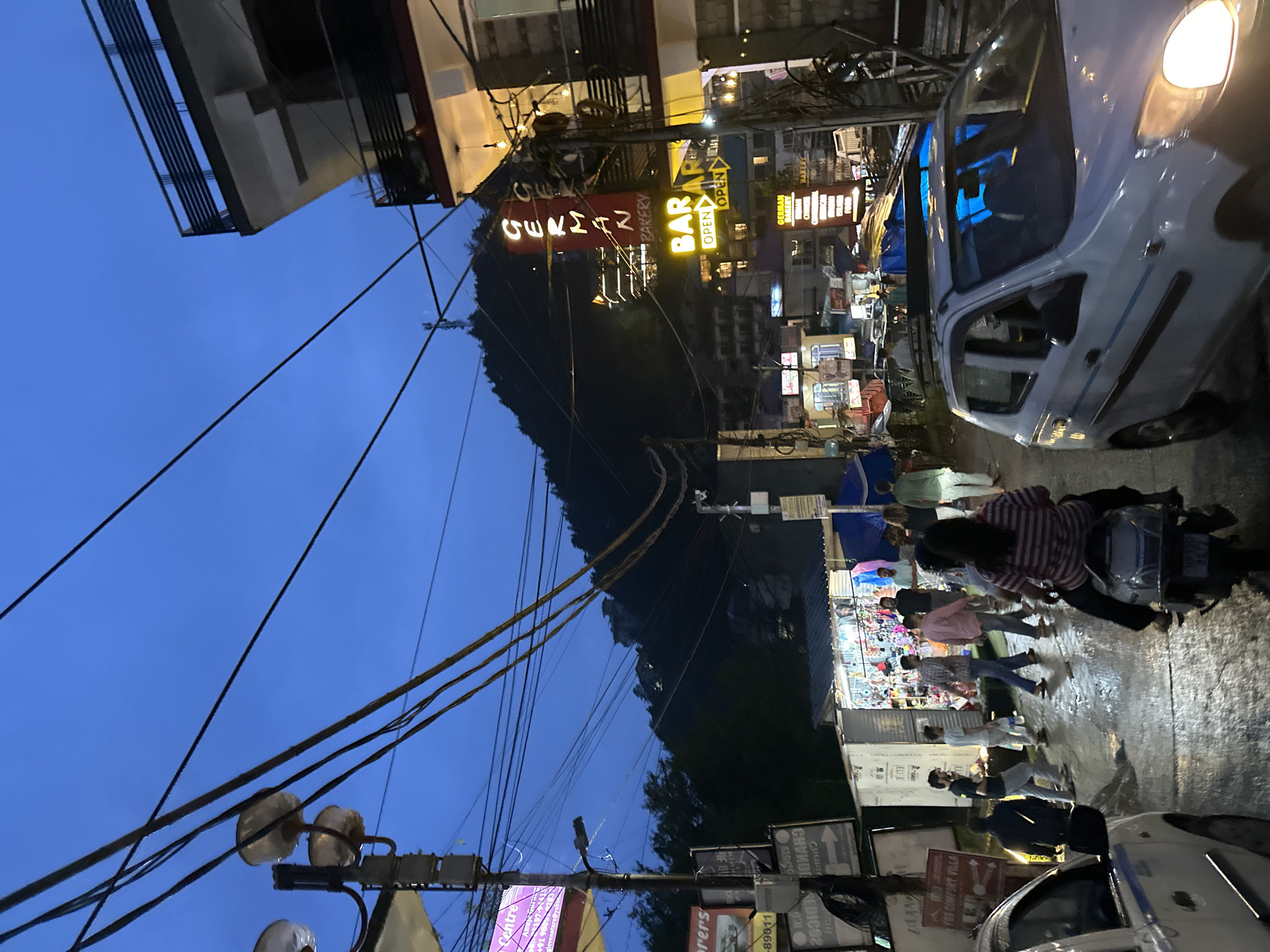 The image depicts a street scene at dusk in an urban environment, likely a town or city, with buildings lining both sides of a wet, paved road. The sky is a deep blue, indicating twilight. Several streetlights are visible, some are on, casting a warm glow. A prominent sign with red lettering reads "GERMAN BAKERY," with a smaller yellow sign below it saying "BAR OPEN." Another sign nearby indicates "BAR."
On the left side of the street, there are various shops and stalls. One stall is open, displaying a variety of goods, with a few people browsing. A white car is parked on the left, and another white car is moving on the right side of the road. A person is riding a scooter in the foreground, facing away from the camera. Several other individuals are walking on the street and sidewalks. Utility poles with numerous wires crisscross the sky, a common sight in many urban areas. In the background, a densely wooded hill rises, with buildings visible on its slopes. The overall atmosphere is that of a busy street at the end of the day, with commercial activity continuing into the evening.