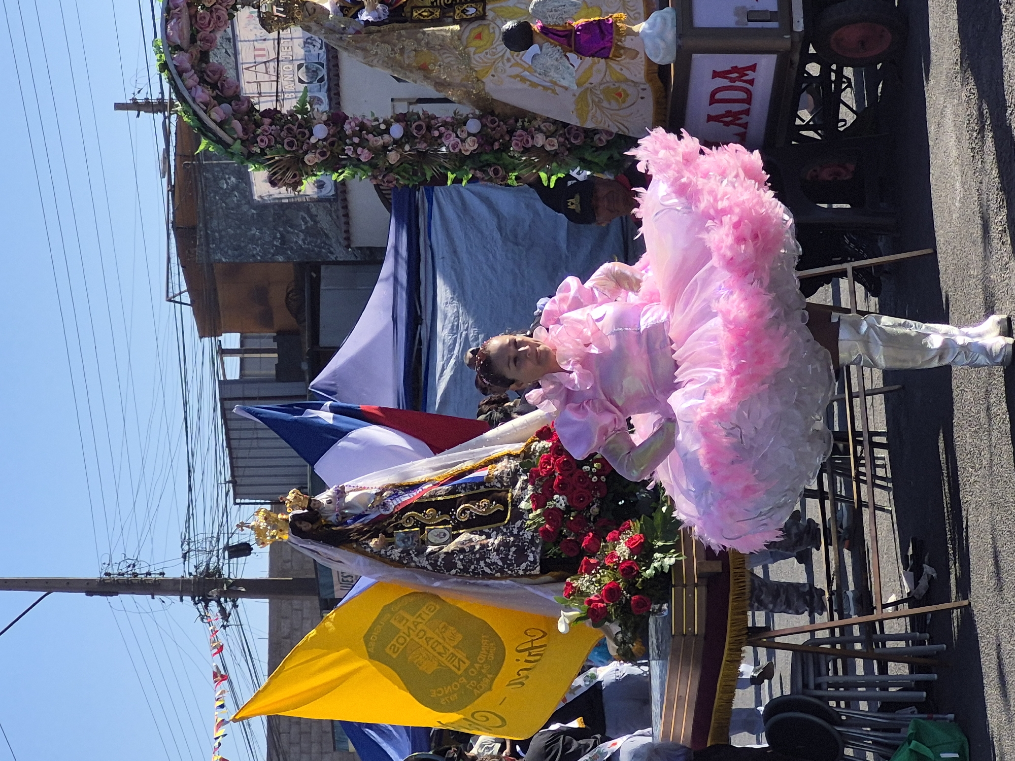 The image depicts a street scene, likely in Arica, Chile, during a religious procession or festival. In the foreground, a woman is dressed in a voluminous, bright pink, ruffled costume with metallic silver boots. She is positioned to the right of a decorated altar or float featuring a statue of the Virgin Mary adorned with a crown and veil, surrounded by red roses. To the left of the statue, a large yellow flag with insignia and text, possibly in Spanish, is prominently displayed. Behind the main subjects, several tents are visible, along with more flags, one appearing to be the Chilean flag. In the background, buildings and a utility pole with numerous wires contribute to the urban setting. A cart with text reading "LADA" in red letters is also part of the scene. The overall atmosphere suggests a public celebration under a clear blue sky.