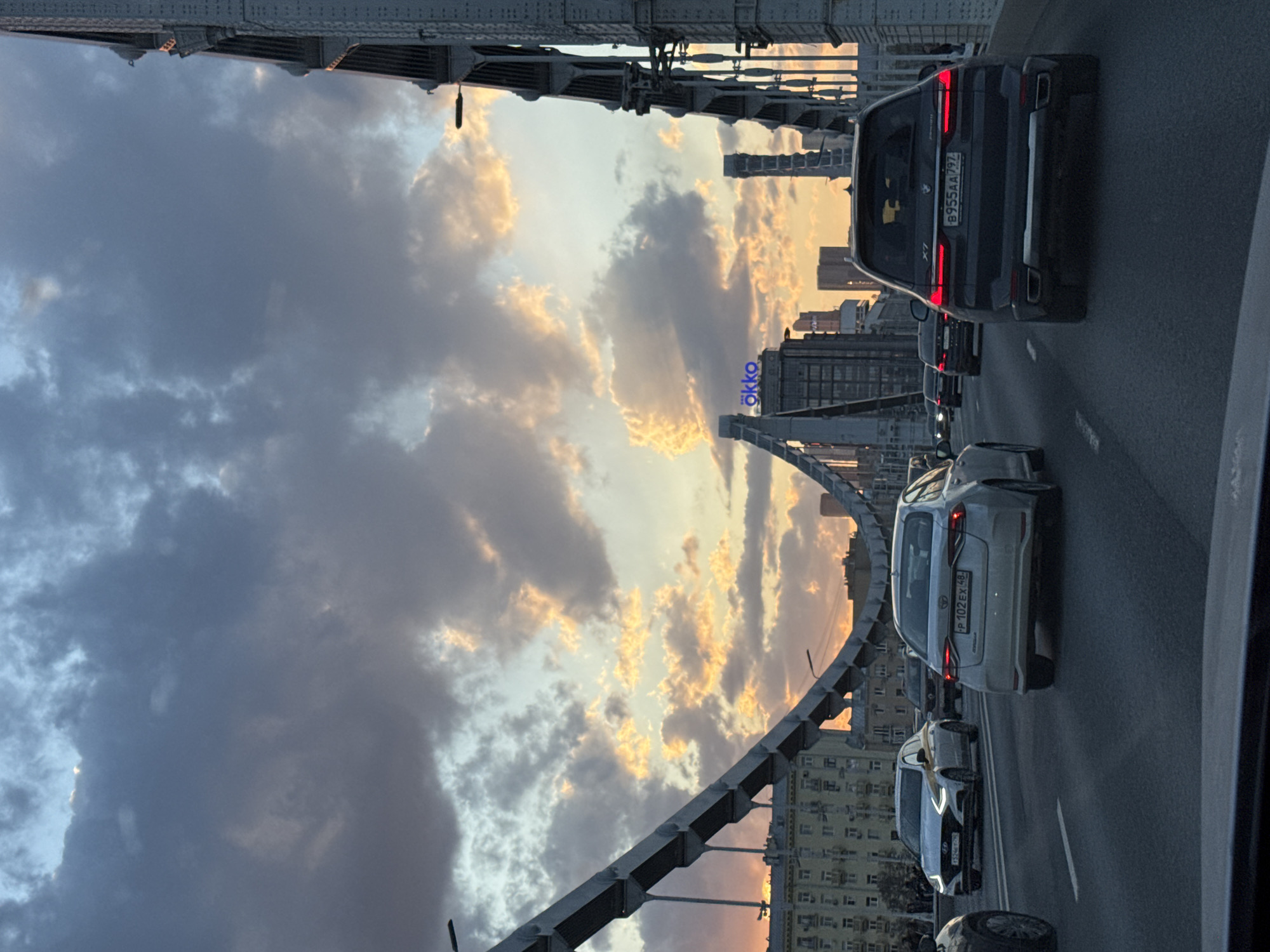 The image shows a view of a roadway in Moscow, Russia, captured from within a vehicle moving along a curved bridge. The perspective is looking forward, capturing the traffic ahead, the sky, and architectural elements of the city.
In the foreground, vehicles are visible in the adjacent lanes. The rear of a dark-colored BMW X7 with license plate "B955AA797" is closest to the camera. Further down the lane, there is a silver car with license plate "P 102 EX 48" and a white car with a black roof ahead of that.
The view also encompasses the metal arches of the bridge. Beyond the arches, there's a view of the sky with dramatic clouds, colored in hues of grey and gold by the setting sun. Additionally, the city's skyline is visible with several buildings. Most notably, the "OKKO" signage is visible on one of the buildings.
The image depicts a typical urban scene during the late afternoon or early evening, with vehicular traffic on a major bridge in Moscow.