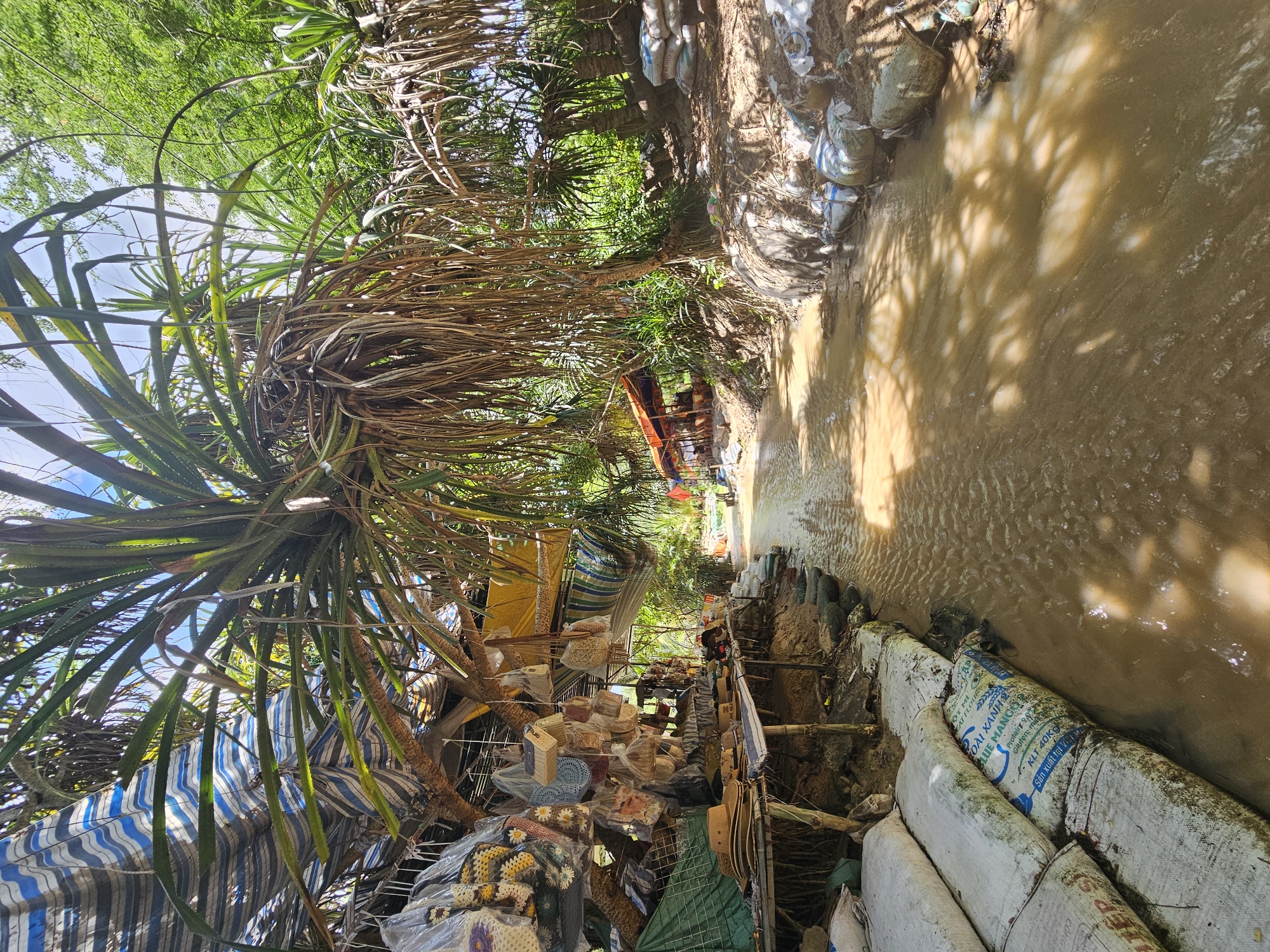 This image captures a scene in Mũi Né, Vietnam, likely a riverside market. The foreground is dominated by a narrow, muddy waterway with reflections of sunlight and shadows from overhead foliage. The banks of the waterway are lined with sandbags. To the left, a makeshift market stall is set up under a canopy of blue and white striped fabric and palm fronds. Various handmade items, including woven bags, hats, and decorative objects, are displayed, many enclosed in plastic packaging. Some of these items appear to be crochet work. A table displays straw hats. Further down the waterway, a small, orange-roofed structure is visible, suggesting a food stall or seating area for customers. The background is filled with dense green vegetation, indicating a natural, possibly rural or semi-rural, environment. The overall impression is one of a humble, tropical market setting by a waterway.