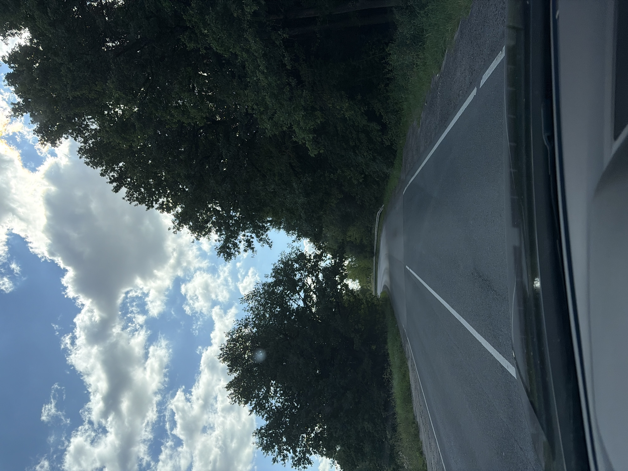The image captures a road curving through a densely wooded area under a partly cloudy sky. The road, paved and marked with white lines, leads into the distance and appears to be a two-lane highway. Tall, dark green trees line both sides of the road, creating a tunnel-like effect. The sky above is a mix of blue and white, with fluffy clouds that cast shadows on the landscape. The foreground shows a portion of a car's interior, likely the dashboard and windshield, suggesting the photo was taken from a moving vehicle. There are no people or animals visible in the scene. The environment appears to be rural and natural, consistent with a location in Kolontaevo, Russia, known for its natural landscapes.