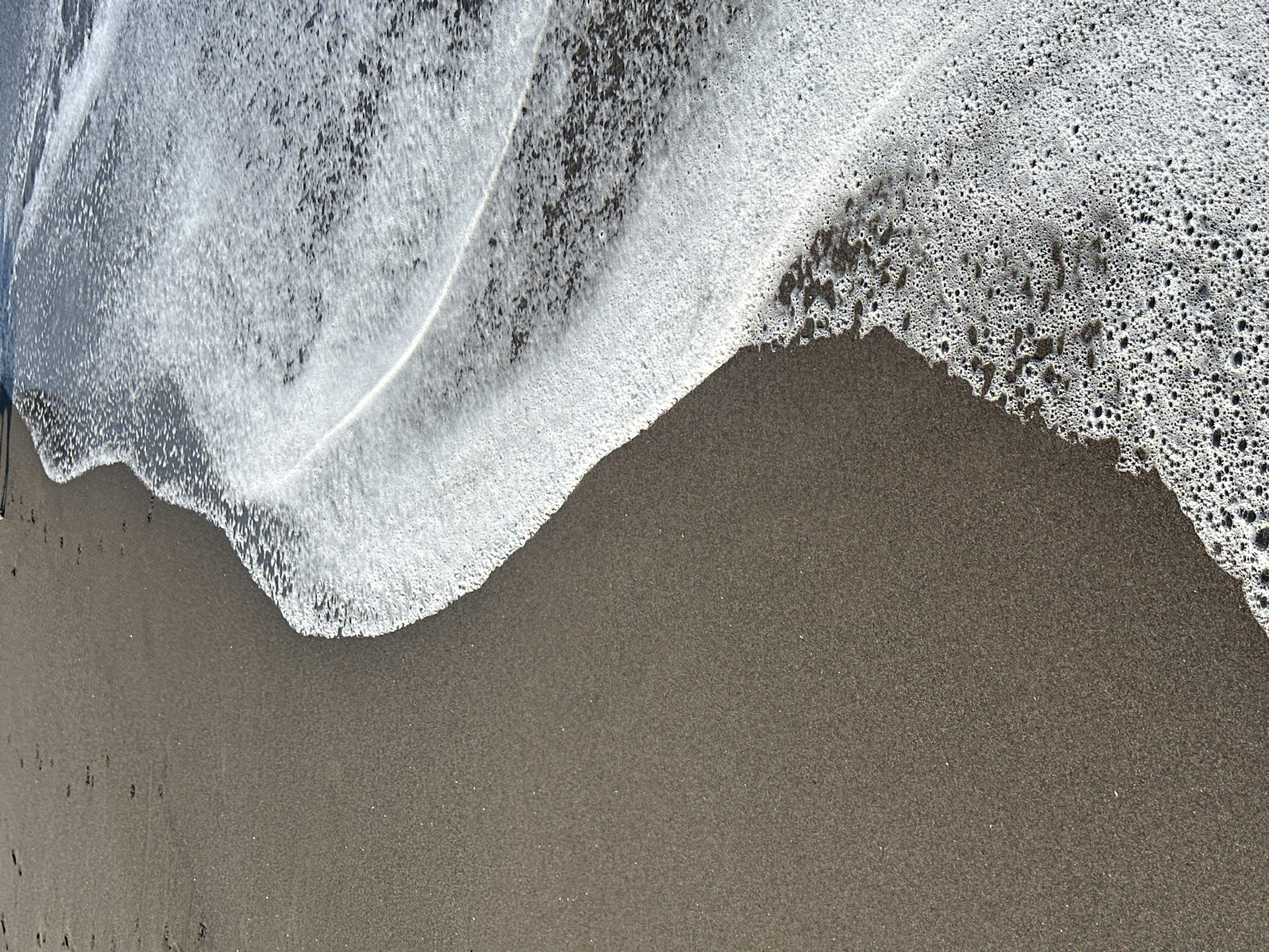 The image shows a close-up view of a sandy beach with a gentle wave receding. The sand is dark brown and appears wet in places. Footprints are faintly visible on the dry sand. The wave has left a line of white foam and bubbles along the shoreline, which is curving from the lower right corner towards the upper left. The water is bright and reflective, creating a shimmering effect on the foam. The scene suggests a tranquil moment at the beach. The image is from North Kuta, Indonesia.
