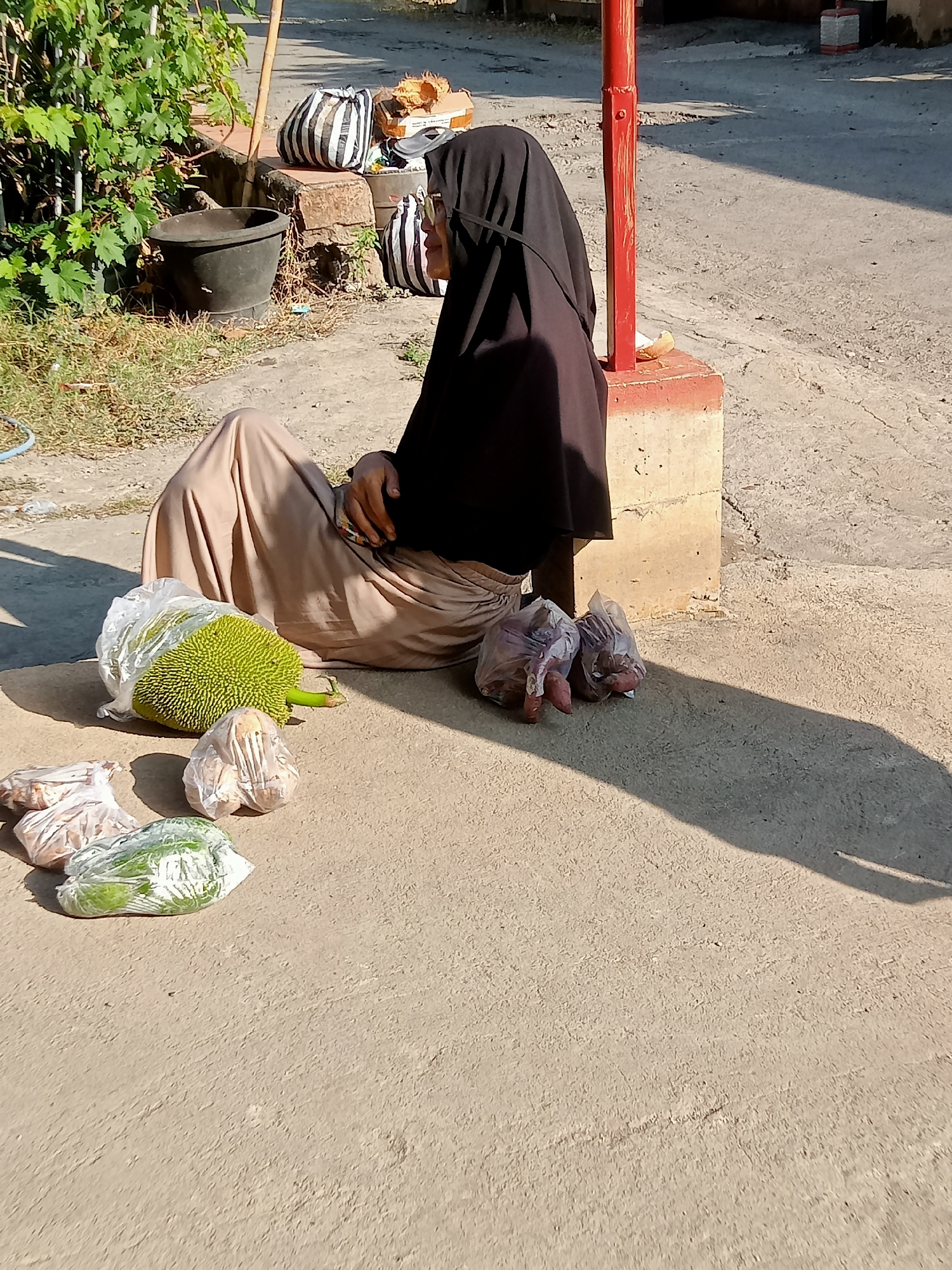 In Simo, Indonesia, a person wearing a black hijab and a tan skirt sits on the ground in front of a red post. The person is facing right, with their left arm resting on their lap. In front of them, several items are placed on the ground in plastic bags: a large green jackfruit, some onions, and some green vegetables. In the background, there is a black pot, some striped bags, and a wooden pole. The ground is paved with concrete and appears cracked in places. The scene is lit by sunlight, casting a long shadow from the red post onto the ground.