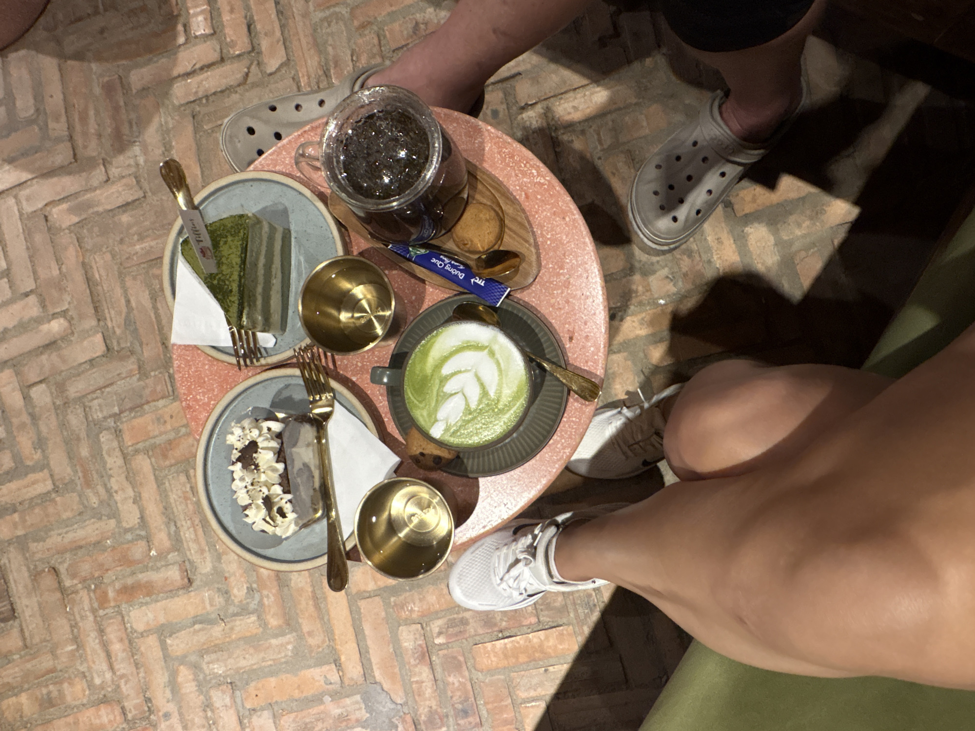 From an overhead perspective, the image shows a small, round, pink table situated on a herringbone brick floor. Two people are visible, indicated by their legs and feet. One person is wearing grey Crocs, while the other wears white sneakers. On the table are two plates of cake, one with green cake and another with chocolate cake, each accompanied by a golden fork and a small golden cup. A grey teacup with a latte art design is also present, along with a golden spoon and a small packet of sugar. A glass teapot filled with liquid and herbs rests on a small wooden tray. The location is suggested to be in or near Bac Natural Heritage Area Trang, Vietnam.