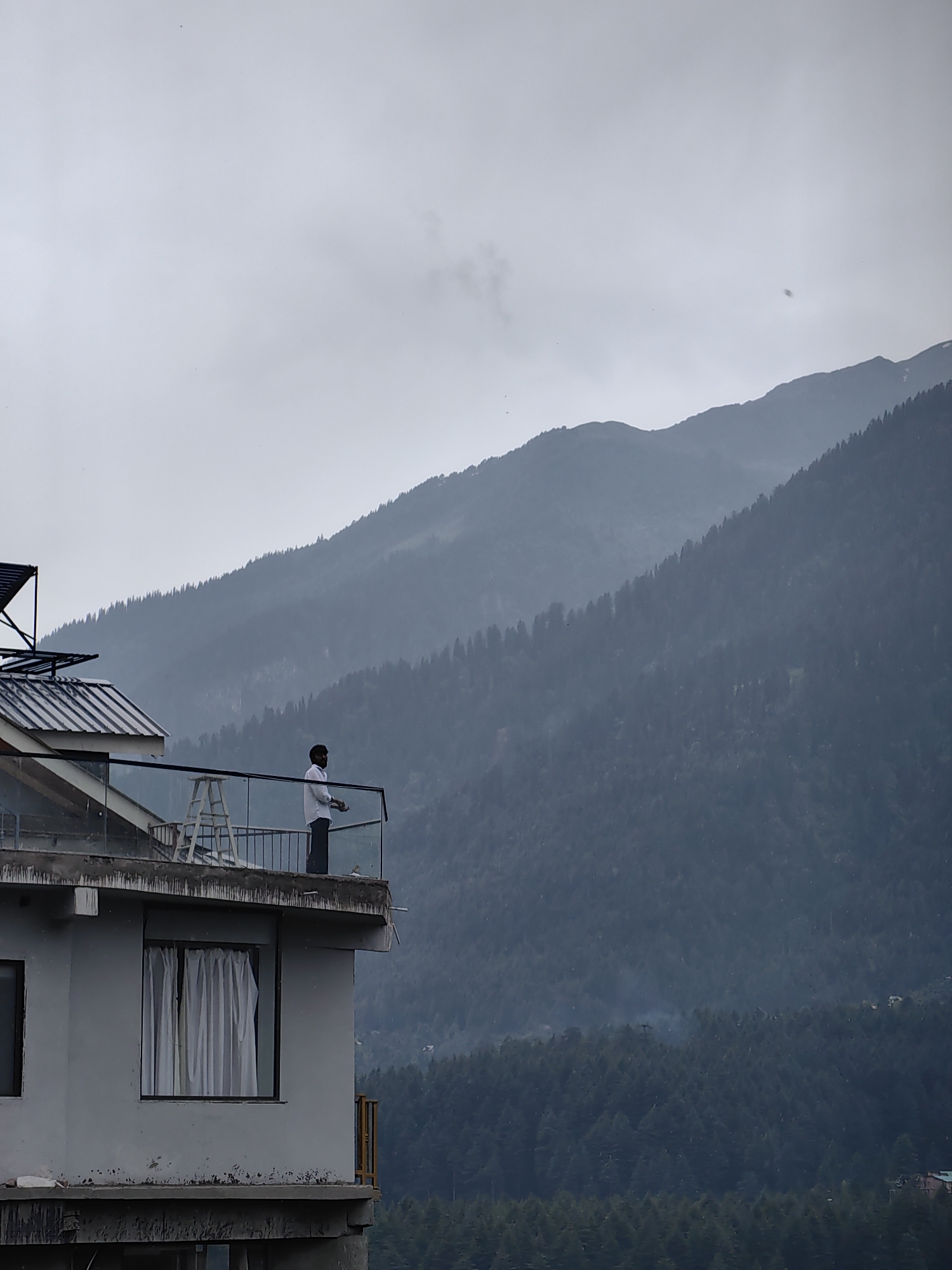 An overcast day features a man in a white shirt and dark pants standing on a balcony of a modern building with a glass railing. The balcony overlooks a dense, dark green coniferous forest that covers the slopes of layered, misty mountains. The building is light grey with a dark-tinted window covered by white curtains on the visible facade. A silver ladder stands on the balcony. Tiny specks, possibly birds or dust, are visible in the sky. The scene suggests a location in Vashisht, India, known for its mountainous terrain and dense forests.