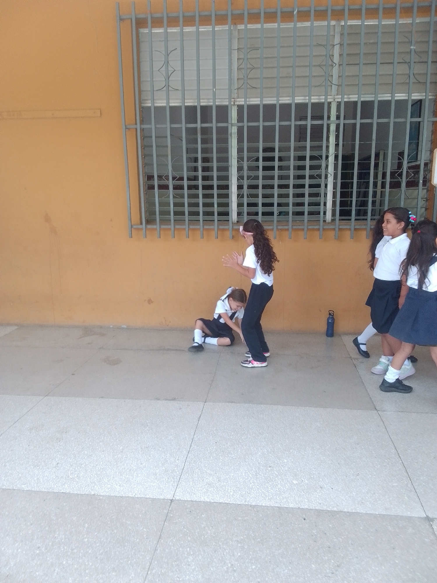 The image shows a group of young girls in school uniforms standing and playing on a tiled walkway outside a building with an orange wall and a barred window. One girl in the foreground, on the right, wears a white polo shirt and a dark skirt with white socks and black shoes. She is looking to her left with a slight smile. Another girl next to her, slightly behind and to her left, is also wearing a white polo shirt and a dark skirt, and appears to be walking forward.
Further to the left, a girl with long curly dark hair and a white shirt and dark pants is looking down with her hands in front of her. She is wearing athletic shoes. To her left and slightly behind her, a younger girl is sitting on the ground wearing a white shirt, dark skirt, white socks, and black shoes. She is looking down at the ground.
A blue water bottle is visible on the ground near the standing girls. The scene appears to be outdoors, possibly during the daytime, given the lighting. The overall atmosphere seems playful and casual, with the children engaged in some sort of game or activity. The tiled walkway and the architecture suggest a school setting, possibly in a warm climate. The location context is La Tagua, Venezuela. There is no visible text in the image.