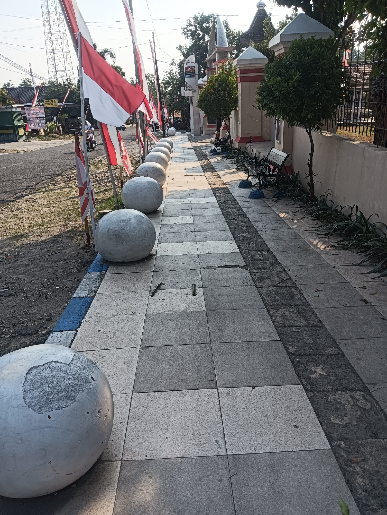 The image depicts a sunny street scene in Palemrejo, Indonesia, with a focus on a sidewalk decorated with several large, round, white concrete planters. Indonesian flags, red and white, are prominently displayed along the sidewalk, attached to metal poles. The sidewalk itself is paved with square tiles in varying shades of gray and white. To the left of the sidewalk is a paved road with a dirt shoulder. A person on a scooter is visible on the road, heading away from the camera. Behind the sidewalk, a beige wall with red accents runs along the property line. Several trees and benches are also present along the sidewalk, suggesting a public or commercial area. Buildings and a tall communication tower are visible in the background. One of the concrete planters in the foreground shows visible damage, with a chunk missing from its surface.
