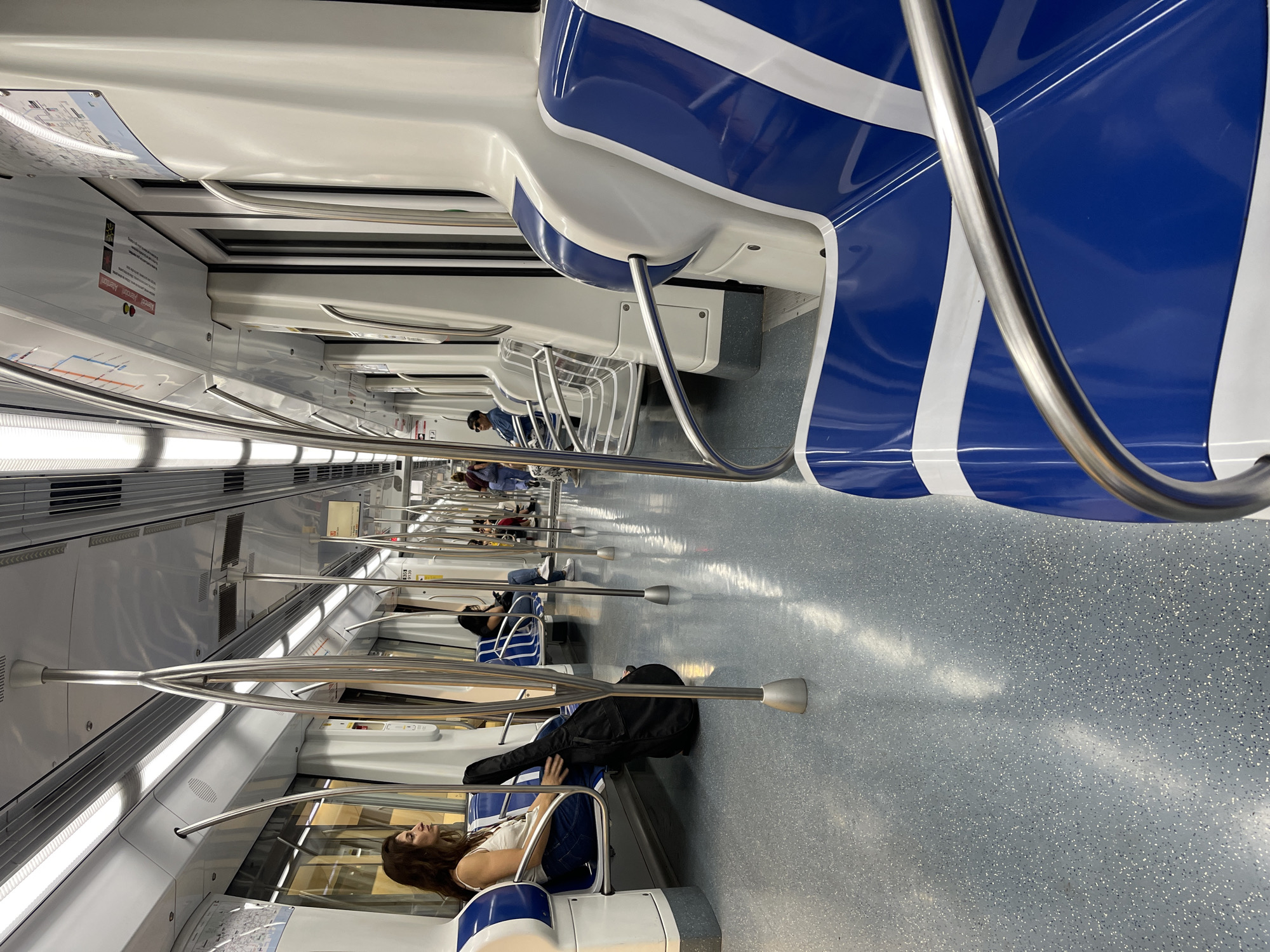 The image depicts the interior of a modern, clean metro train car in Barcelona, Spain. The seating is predominantly blue and white, with a metallic pole system for passengers to hold onto. The floor is a light grey speckled material. The lighting is bright and fluorescent, originating from long fixtures on the ceiling.
In the foreground, to the right, a close-up view of a blue and white seat with a chrome handrail is visible. Further into the train car, a woman with long, dark, wavy hair is seated on the left, wearing a white top and blue jeans. Next to her is a black instrument case, likely for a guitar. She is looking towards the left side of the train car. In the background, other passengers are sparsely seated, with one individual wearing sunglasses and a denim shirt to the left and others further down the aisle. The overall impression is one of calm and order within the public transportation system.