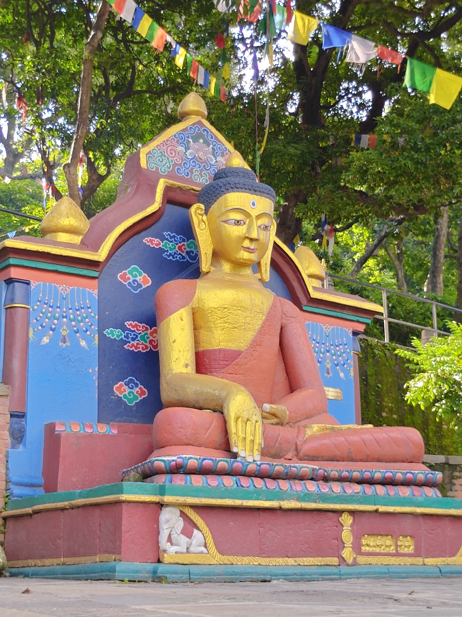 The image shows a seated, golden Buddha statue, adorned with a red robe. The statue is part of a vibrantly decorated shrine, featuring blue, red, and gold accents with intricate cloud and floral motifs. Prayer flags in various colors (white, blue, red, green, yellow) are strung across the top of the frame, suggesting a spiritual or religious setting, likely within the Kathmandu Metropolitan City, Nepal. The background consists of lush green trees and foliage, indicating an outdoor location. The statue is positioned on a multi-tiered, colorful pedestal decorated with further symbolic carvings, including an elephant motif on the lower section. The lighting suggests daylight, with some sunlight filtering through the trees.