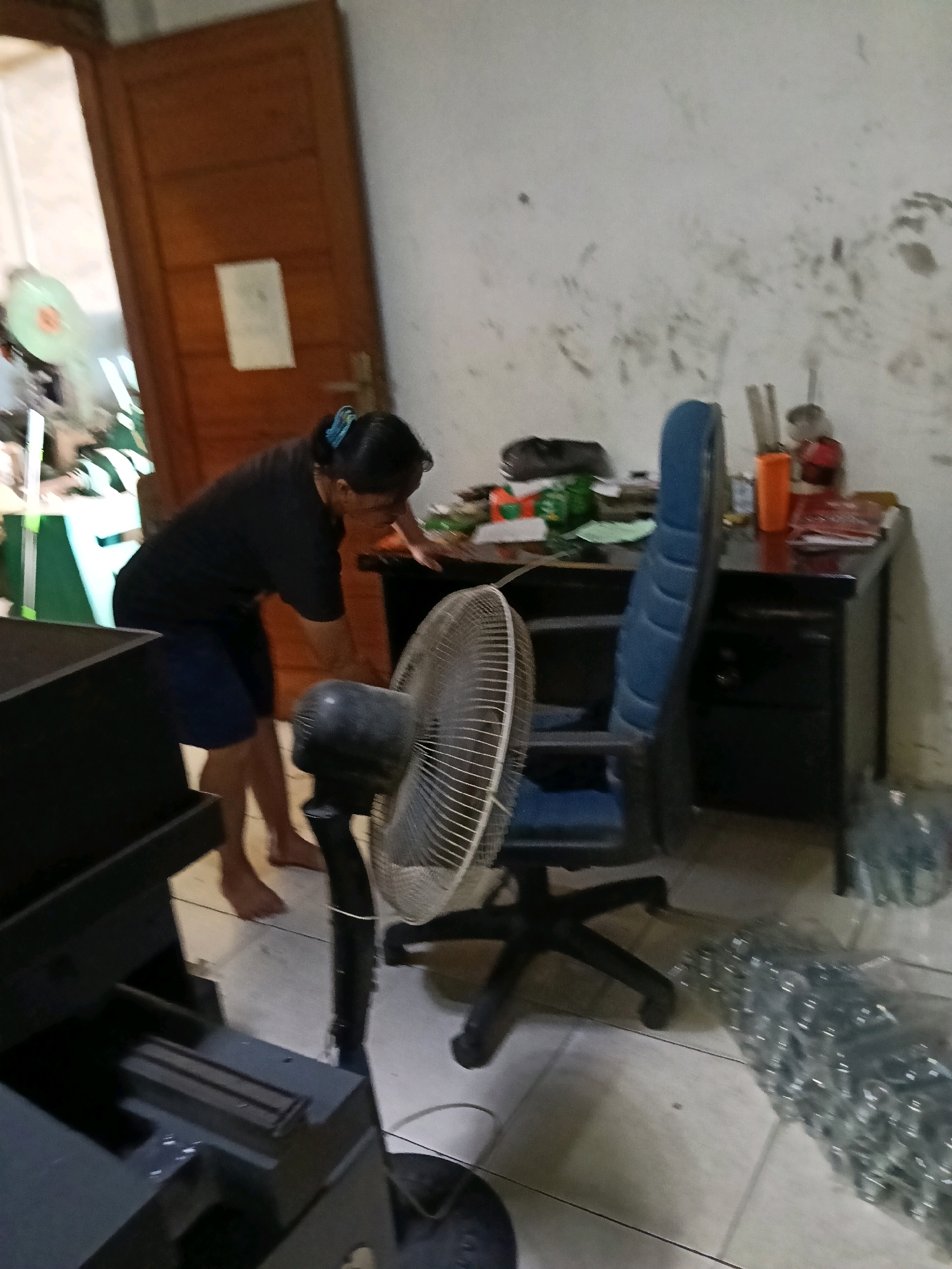 A person is bent over a desk, reaching for an object. The person is wearing a black t-shirt and blue shorts. They have their hair tied back with a blue hair tie. In the foreground, a large fan is turned on. Behind the desk, there is a blue office chair and a wall with scuff marks. To the left of the desk, there is a dark colored cabinet. To the right of the desk, there are stacks of clear plastic bags filled with small, metallic objects. The flooring is tiled. The scene appears to be indoors, possibly a workshop or storage area in the city of Belimbing, Indonesia.