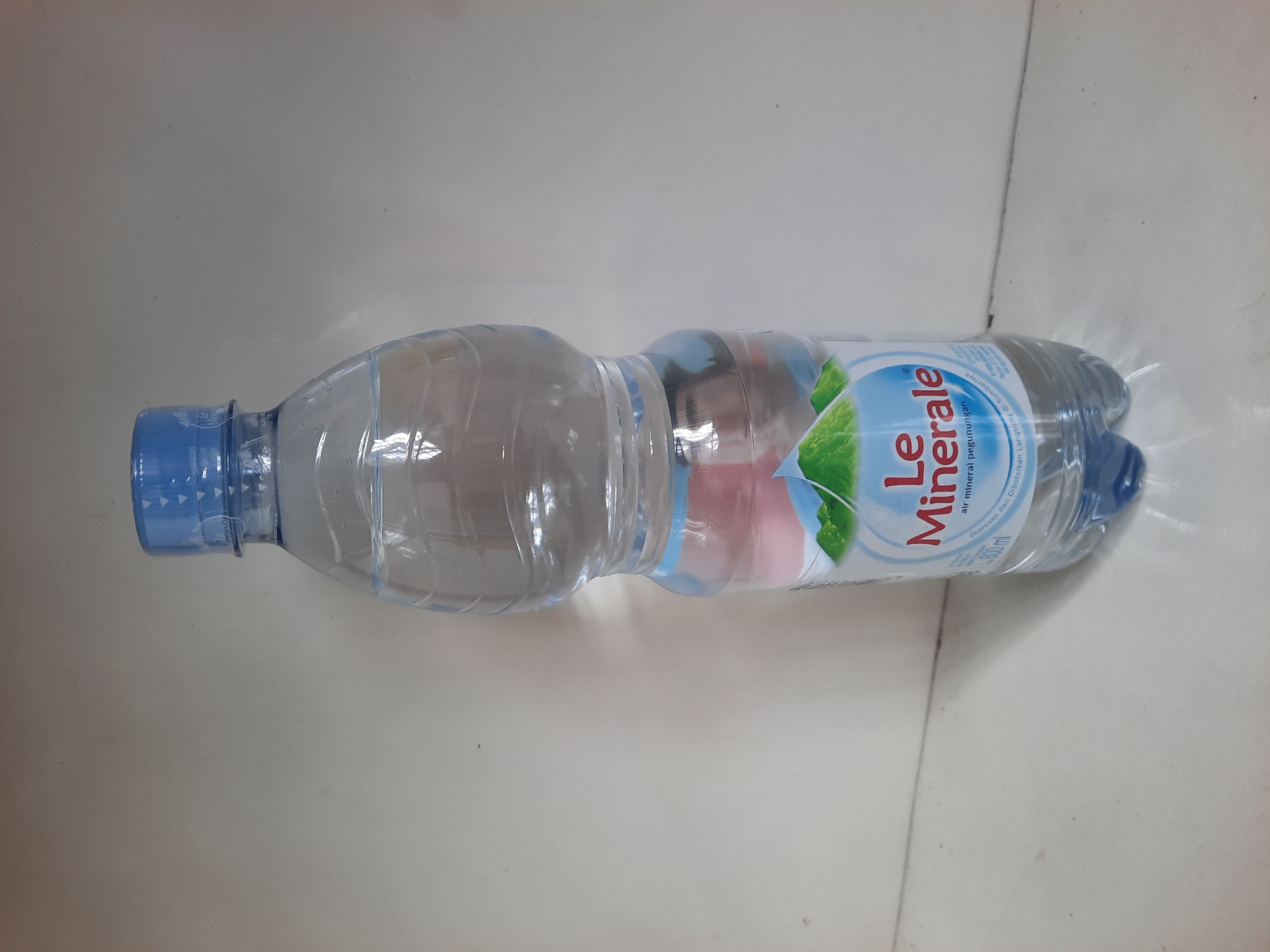 A plastic bottle of Le Minerale bottled water is upright and centered against a plain, light-colored background. The bottle is approximately three-quarters full of clear liquid. The label on the bottle is visible, featuring the "Le Minerale" logo, an image of green mountains, and text in Indonesian. The bottle has a blue plastic cap and a clear plastic body. The surface on which the bottle rests is also light-colored and appears to be flat. The background is a simple, undecorated wall. The lighting suggests it is indoors, and there are no other objects or people visible in the frame. The image does not provide any specific information about the location in the city of Lhokseumawe, Indonesia.