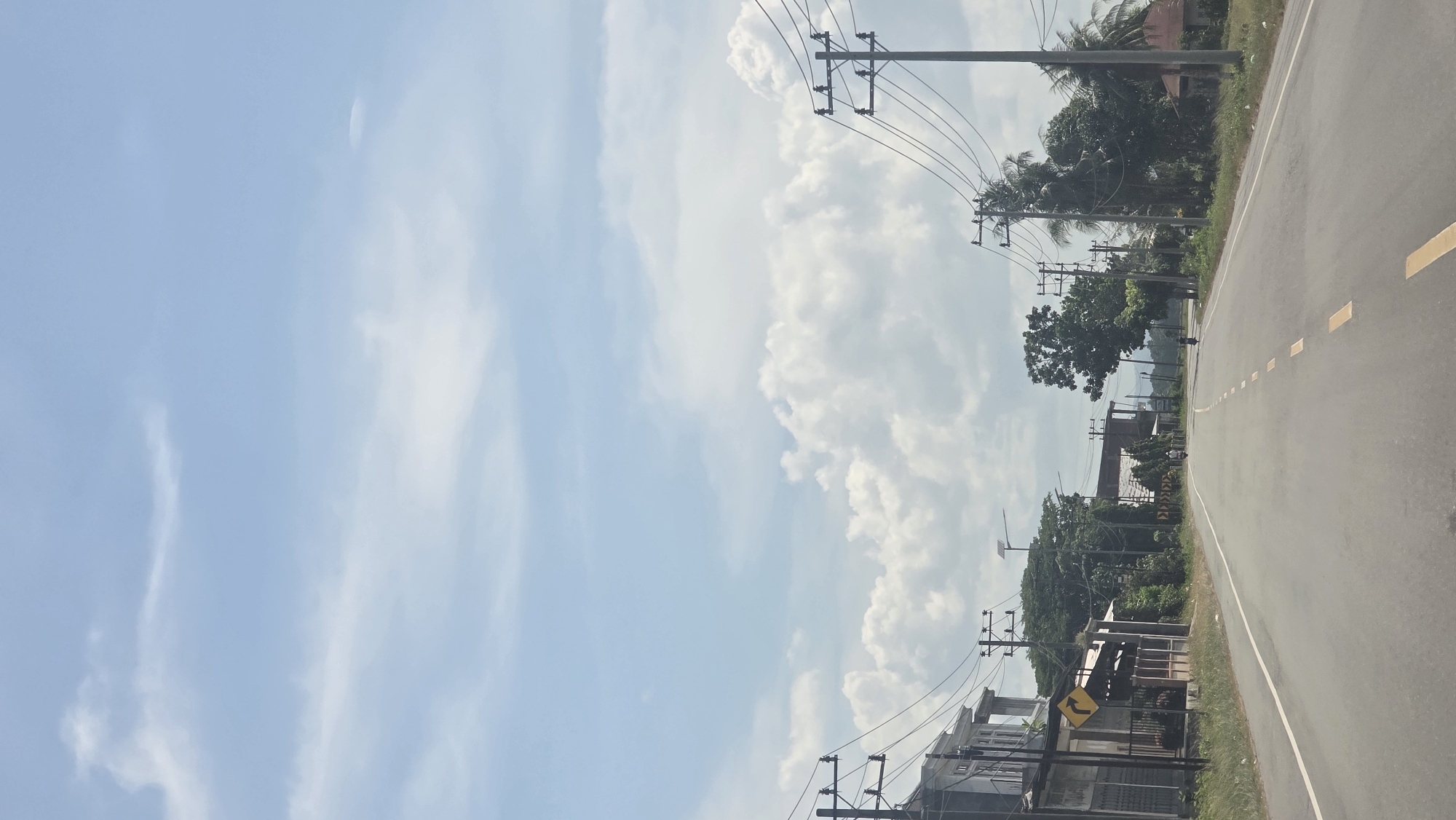 The image shows a road in Jurong Pandee, Indonesia. The road is paved and has yellow dashed lines. Utility poles with wires run along the side of the road, near some trees and houses. The sky is visible with white clouds.