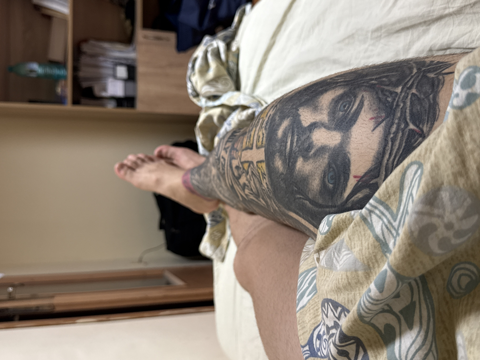 A close-up shot shows a person's lower leg with a large tattoo of a face, possibly religious, with a crown of thorns and red marks resembling blood. The leg is partially covered by patterned fabric, possibly a blanket or clothing. The person's foot is visible, extending towards the left of the frame. The background includes a shelf unit with various items stacked, and a glimpse of a floor. The location and city are unknown.