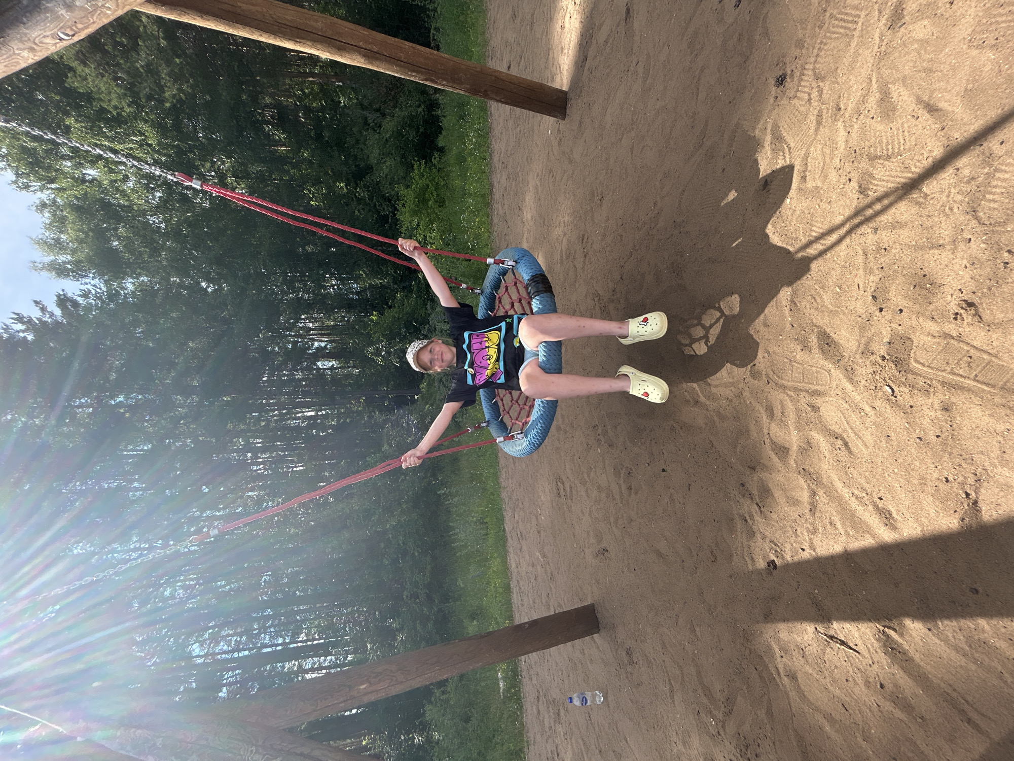 A young girl wearing a black t-shirt with a colorful graphic and light-colored clogs sits on a round blue swing in a sandy area. She is wearing a white baseball cap. The swing is suspended by red ropes and metal chains from a wooden structure, with two thick wooden posts visible. Behind the swing set is a dense forest with tall trees. Sunlight beams through the trees from the upper left, casting a lens flare effect and creating shadows on the sand. A plastic water bottle stands upright on the sand to the left of the swing set. The scene appears to be an outdoor playground in a wooded area, possibly located in Chaikovsky, Russia, given the context.