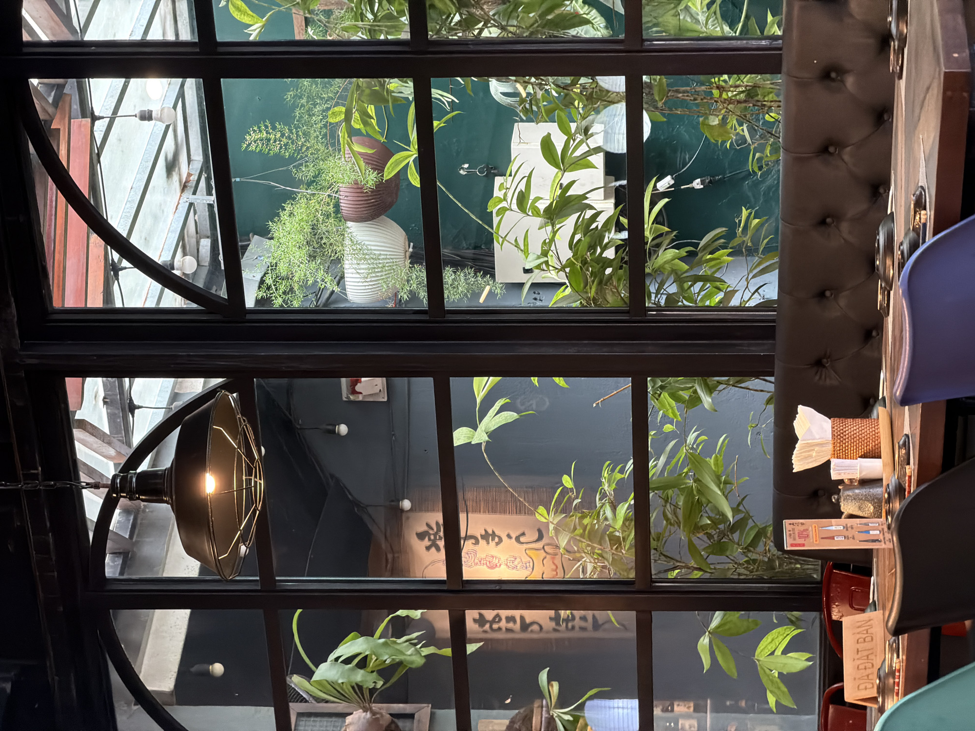 The image provides an upward view from inside a restaurant or cafe, looking through a large, multi-paned window or skylight structure.
**Subjects & Scene:** The interior perspective includes a dark, tufted booth seat on the right and a wooden dining table. Parts of a blue and a red chair are visible around the table. A black, industrial-style hanging lamp with a glowing bulb illuminates the lower-left section. Green foliage is interspersed throughout the frame, both as indoor decor and as part of the outdoor environment.
Through the window, an outdoor or semi-outdoor space is revealed. This area features more green plants, some housed in brown and white ribbed pots. The background consists of building facades or walls in varying shades of dark green, grey, and reddish-brown. Several bare light bulbs, strung on wires, are visible across the exterior, suggesting ambient outdoor lighting. A large, white rectangular object is also noticeable in the outdoor area.
**Activity or event taking place:** The scene depicts a prepared dining table, indicated by the table setting, chairs, and a "TABLE RESERVED" sign. No people are present, suggesting the table is awaiting diners or has been recently vacated. The overall impression is that of a quiet moment in a dining establishment.
**Notable details:** The visible illumination from the indoor lamp and the outdoor bare bulbs suggests it is either evening or night, or an indoor space where artificial light is predominantly used. The combination of green plants, the industrial light fixture, and the outdoor urban elements creates a contemporary, somewhat eclectic ambiance. The location context is Nha Trang Ward, Vietnam.
**Visible text:** On a small wooden sign placed on the table, the Vietnamese text "ĐÃ ĐẶT BÀN" (TABLE RESERVED) is clearly visible. A menu card on the table displays some graphics and small, indistinct text, potentially including "40%". Through one of the windowpanes, a banner or sign with Japanese characters can be seen, specifically "やきとり" (yakitori, meaning grilled chicken skewers) and another phrase that appears to be "ざっか" (zakka, referring to everyday goods or miscellaneous items), suggesting a nearby Japanese eatery or store.