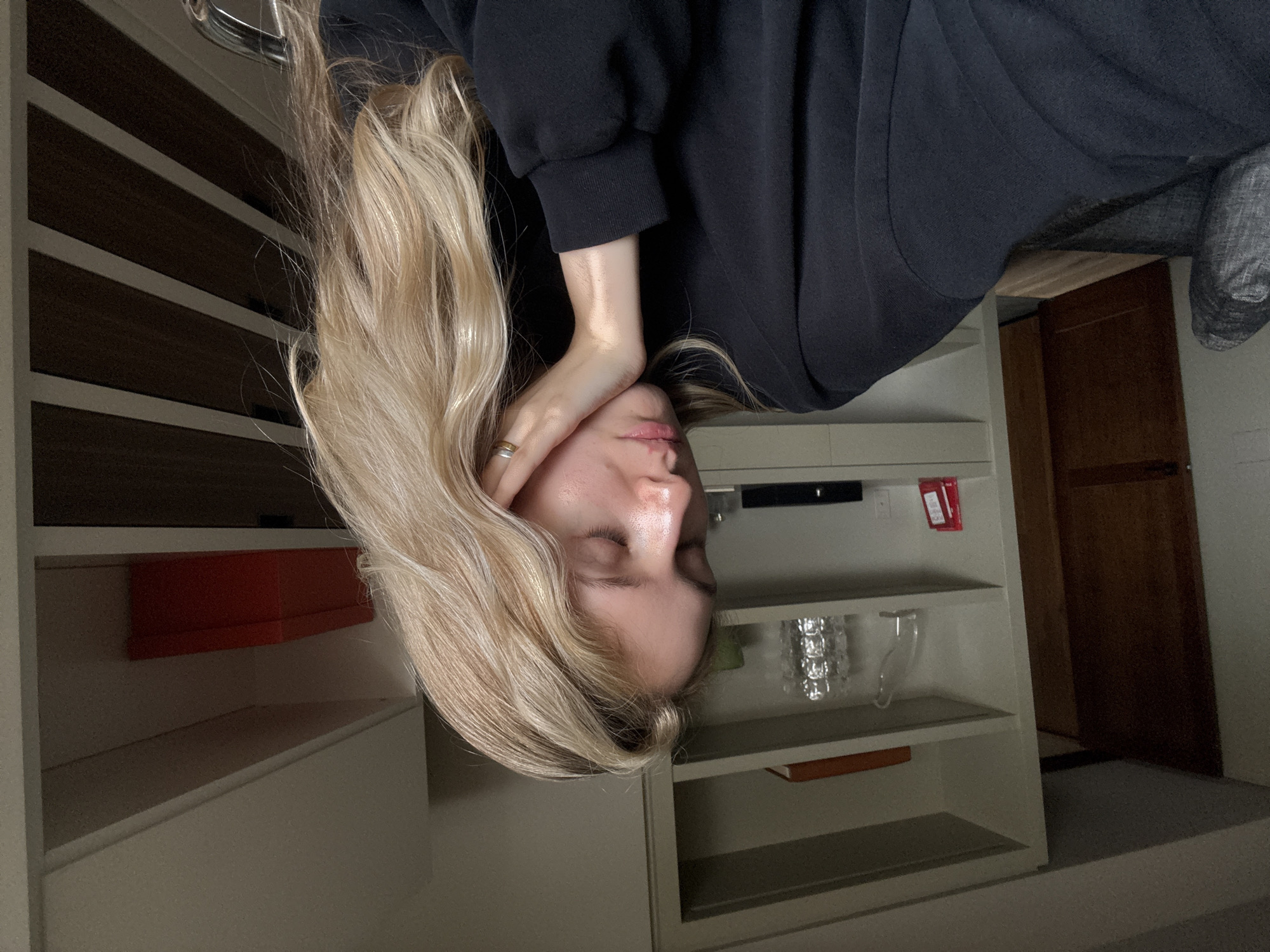 A woman with long, blonde hair and wearing a black hoodie is upside down. She is resting her head on her hand, and her eyes are closed. Behind her is a white shelving unit with various items on the shelves, including an orange box, glassware, and a red object with white writing. A wooden door is visible in the background. The image appears to be taken indoors. Location is not deducible from the image.