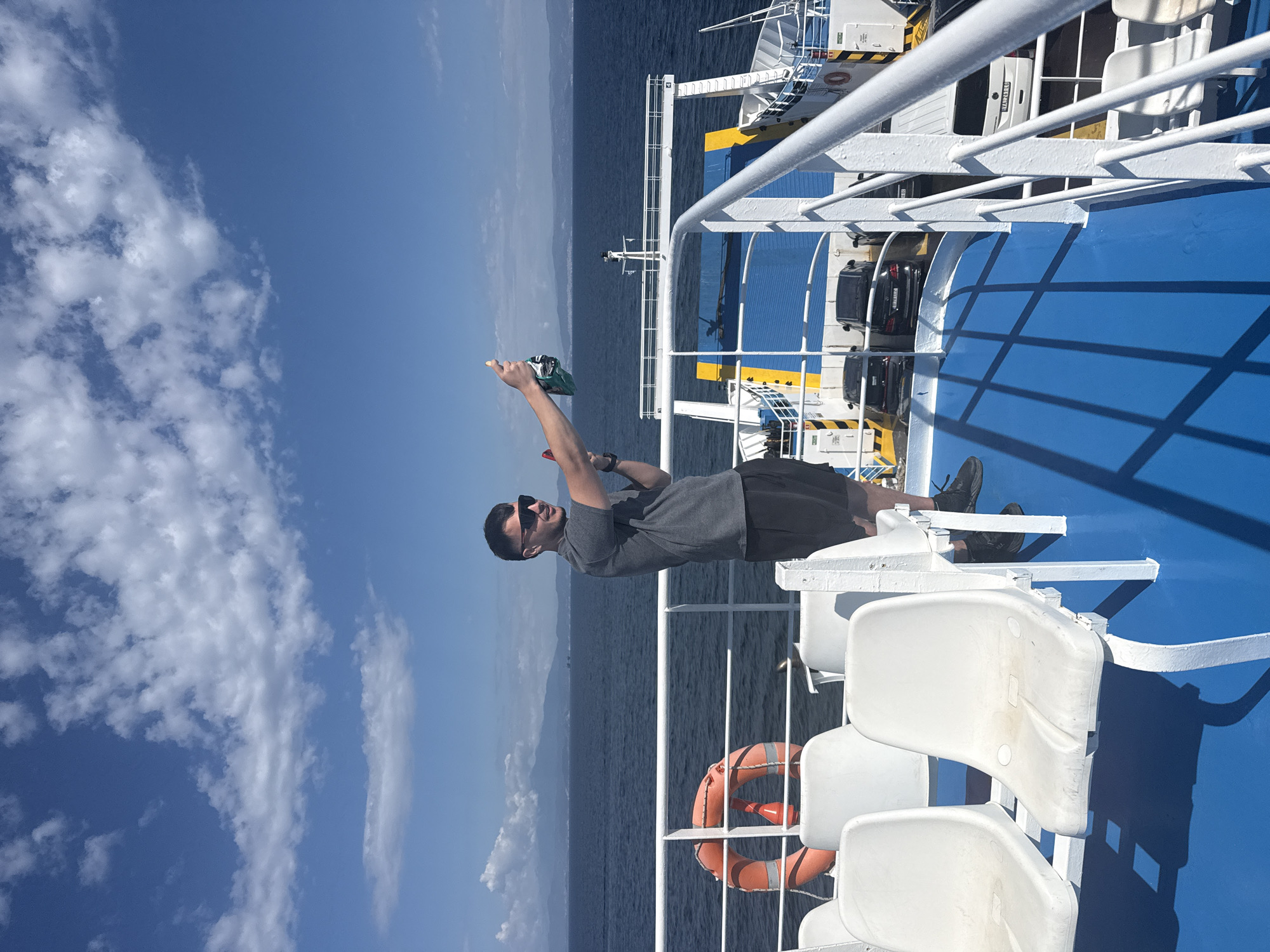 A man stands on the deck of a ferryboat, holding a bottle of champagne and a glass, appearing to take a selfie. He is wearing sunglasses, a grey t-shirt, and black shorts. White plastic seats are arranged in rows behind him. The ferryboat has a bright blue deck and white railings. Beyond the railing, the blue ocean stretches to the horizon, meeting a sky dotted with white clouds. In the distance, a mountainous coastline is visible. The scene appears to be near Prinos, Greece, as indicated by the ferry's operation in that area. The man is captured in mid-action, possibly celebrating or documenting a moment during the ferry ride.