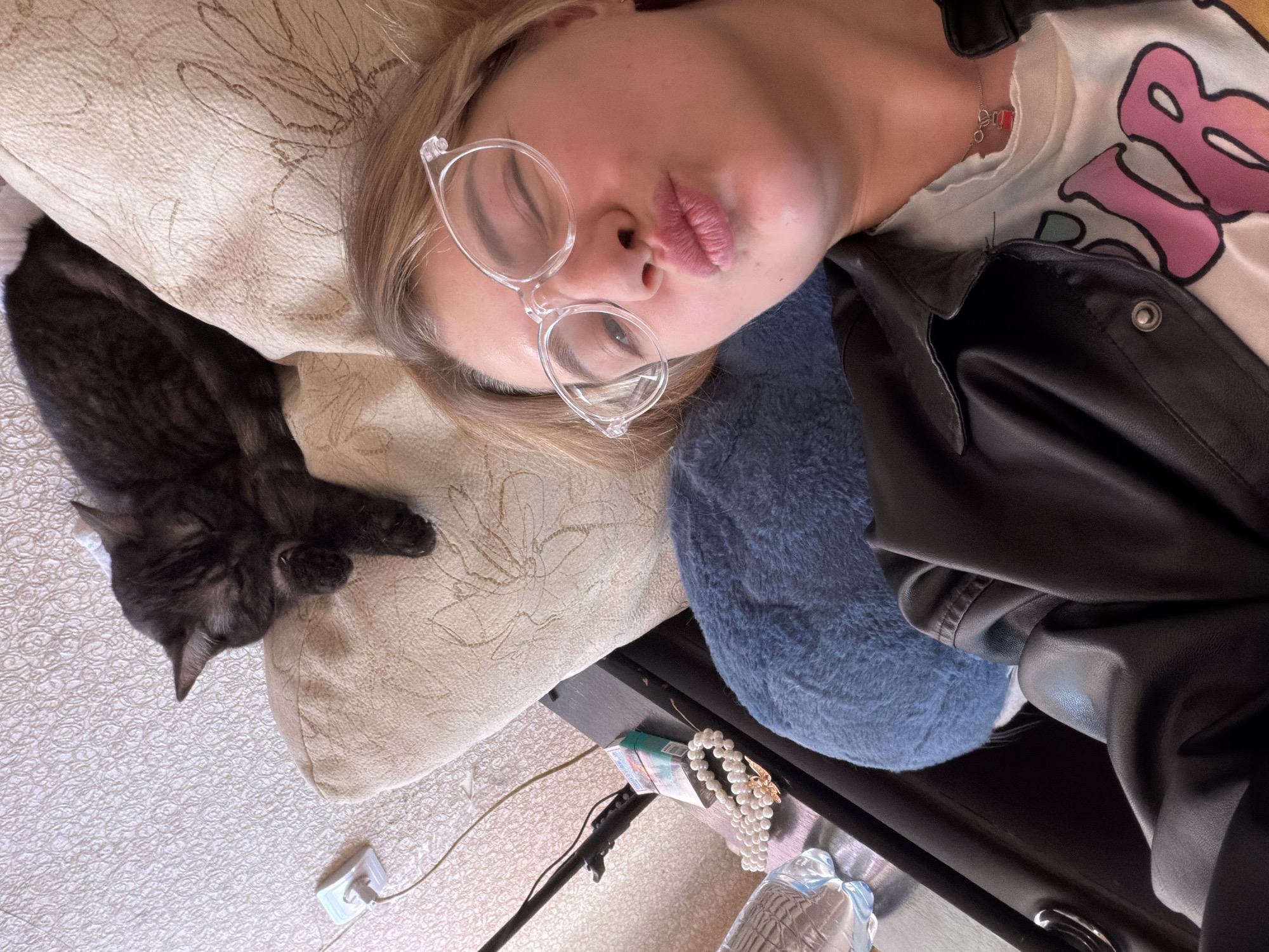 A person lies on a light-colored patterned pillow next to a black cat. They wear clear-rimmed glasses, a white t-shirt with a colorful design, and a black leather jacket. Underneath the pillow, there is a small dark wooden table with a pack of cigarettes, a pearl necklace, and a blue fuzzy object. An electrical plug and wire are on the floor near the pillow. It is impossible to determine the specific location within Naberezhnye Chelny without further information.