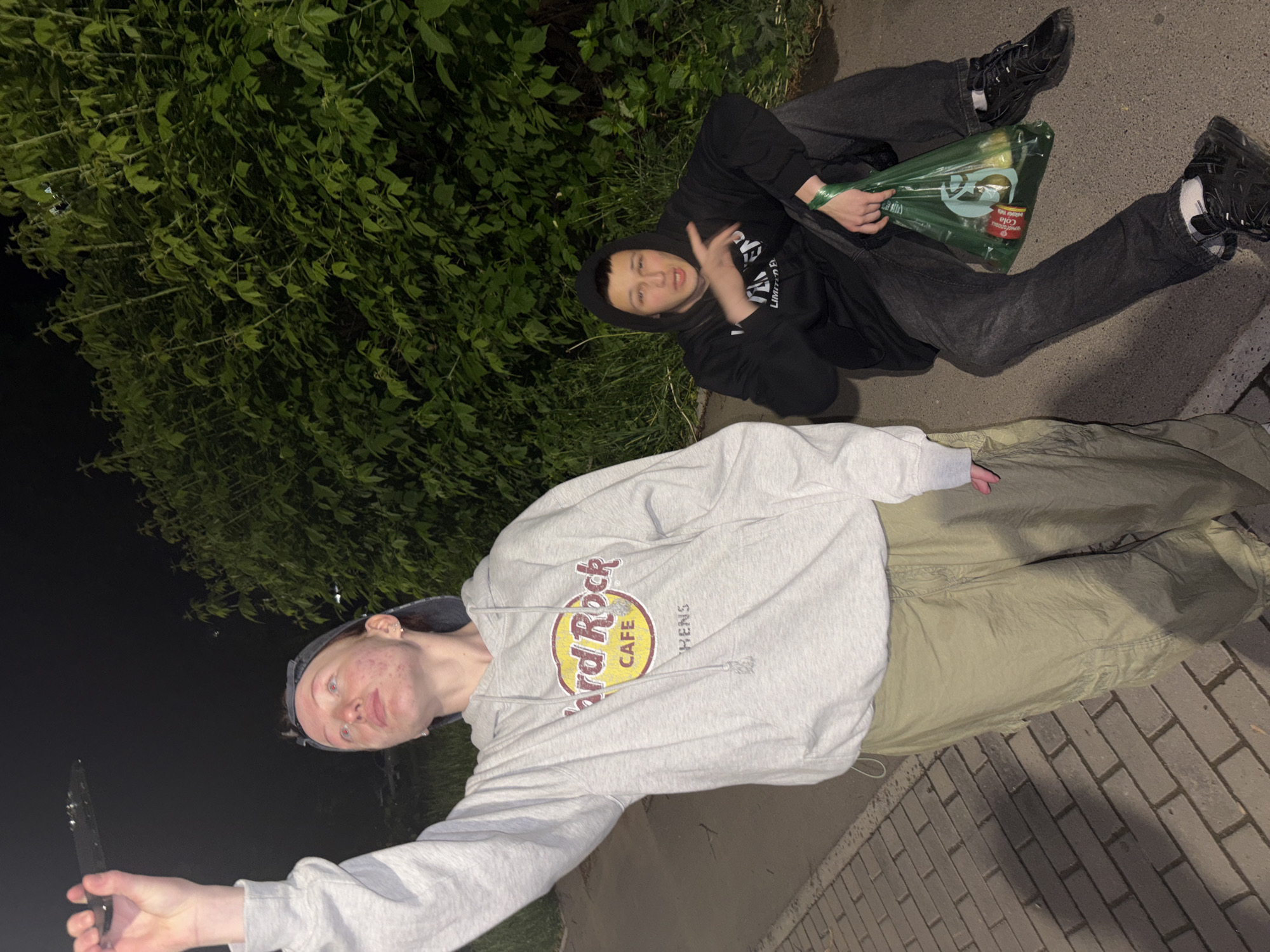 Two individuals are present in an outdoor setting at night. One person, wearing a grey "Hard Rock Cafe Athens" hoodie and light-colored pants, is holding a knife and angled towards the camera. The second person is wearing a black hoodie and dark pants, holding a plastic bag with items that appear to be cola products. The environment includes shrubbery and a paved surface, possibly a sidewalk or walkway. The location is in the city of Chaikovsky, Russia.