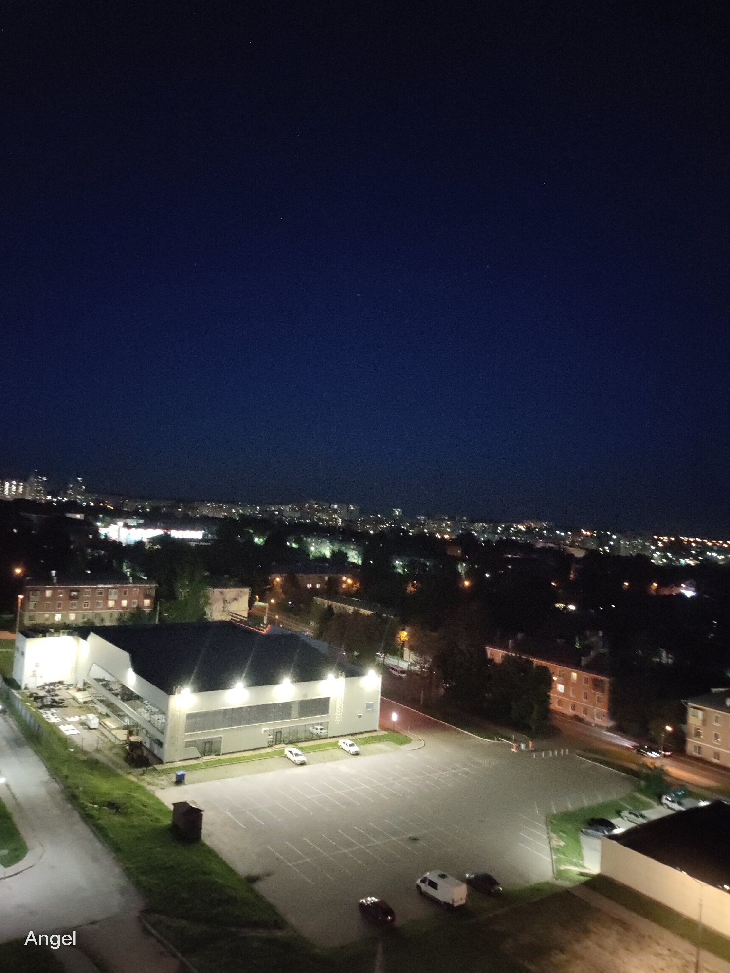 The image captures an aerial view of an urban landscape at night, likely in Tula, Russia, given the provided context. The sky is a deep, clear blue-black, indicating nighttime. In the foreground, a modern, multi-story building with a dark roof and illuminated windows is visible. Adjacent to this building is a large, empty parking lot marked with white lines, with a few vehicles scattered throughout. A small, dark, wooden structure, possibly a shed or storage unit, sits on a grassy patch next to the parking lot.
Further into the midground, there are residential buildings with illuminated windows, suggesting occupied apartments. Trees are scattered throughout this area, their dark foliage contrasting with the artificial lights. A street lamp is visible, casting a warm glow on the surroundings.
In the background, the distant cityscape of Tula is visible, characterized by a dense cluster of buildings and a multitude of lights. The overall impression is one of a quiet urban evening, with the city lights creating a warm glow against the dark night sky. The image appears to be a still photograph, with no discernible movement or action taking place. The quality of the image suggests it was taken with a mobile device or a digital camera with good low-light capabilities. The word "Angel" is visible in the lower-left corner of the image, possibly a watermark or a personal annotation.