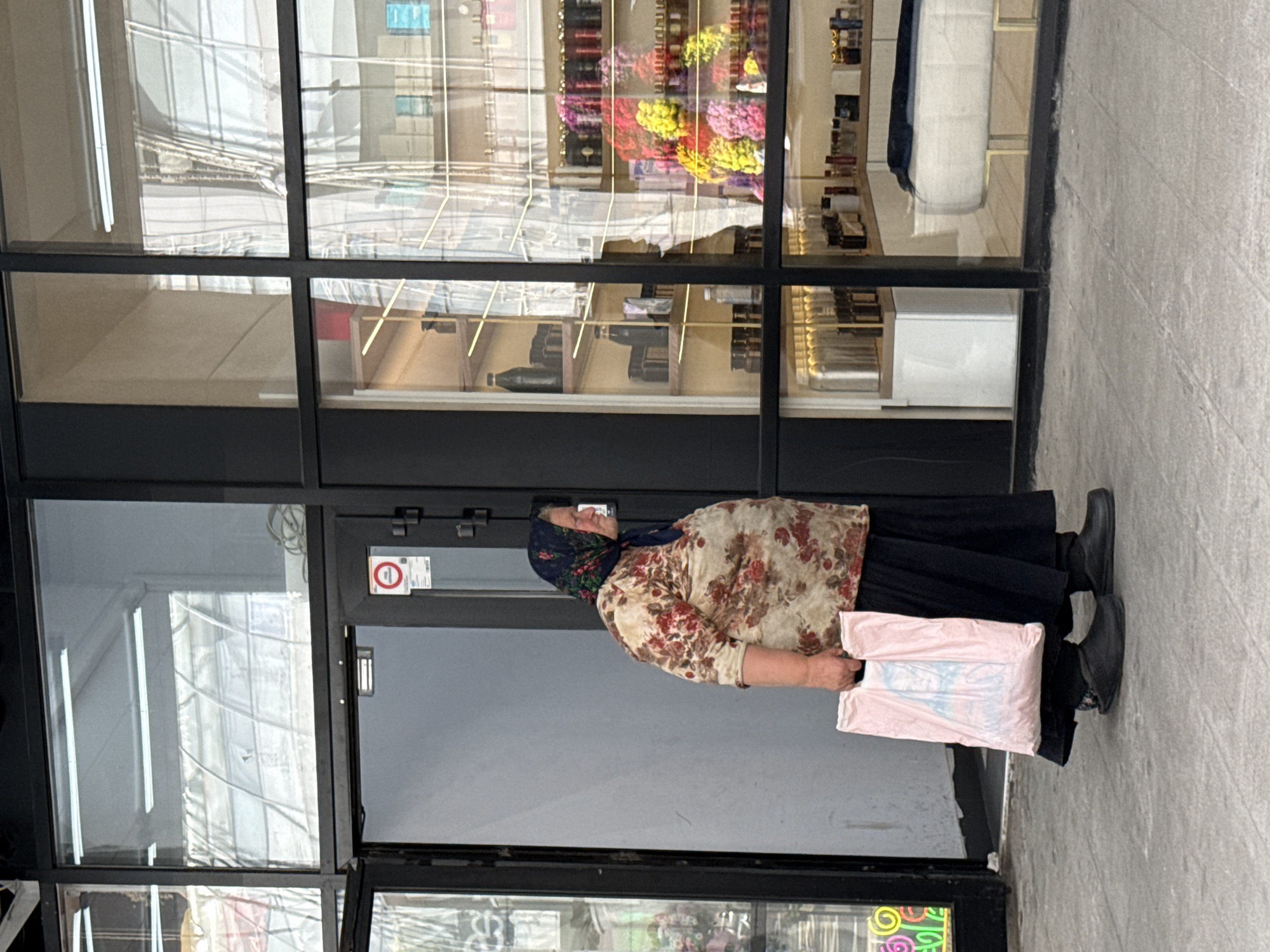 An elderly woman wearing a floral-patterned shirt, a dark headscarf, and a dark skirt walks past a shop window in Moscow, Russia. She is holding a pink plastic bag in her left hand. The shop appears to be a boutique selling perfumes and cosmetics, with rows of bottles and colorful floral arrangements displayed on shelves behind the glass. The woman is walking on a paved sidewalk in front of the shop. The shop entrance is visible on the left side of the image, with a glass door and a sign on the wall. The background shows reflections of buildings and possibly an indoor market or atrium with a curved roof structure. The overall scene depicts a slice of urban life on a day with overcast skies.