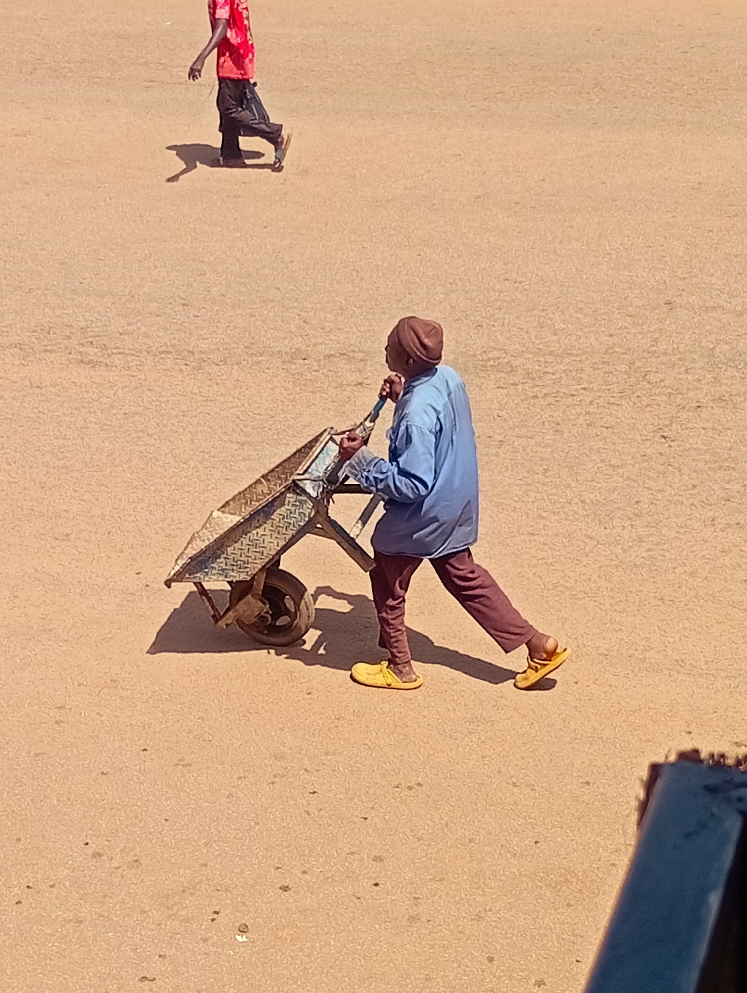 An outdoor, eye-level shot depicts a man pushing a wheelbarrow across a sandy or dirt surface, likely in Bukuru, Nigeria. The man, wearing a brown beanie, a light blue long-sleeved shirt, and dark red trousers, is captured in profile, mid-stride, with his right hand on the wheelbarrow's handle. His yellow sandals are visible. The wheelbarrow itself is metallic with a diamond-plate pattern on its basin and a single wheel. In the background, another person is partially visible, wearing a red shirt and dark trousers, walking away from the camera. The ground is a uniform sandy color with scattered dark specks. A blue object, possibly a railing or part of a structure, is visible in the lower right corner of the frame. The scene is illuminated by bright sunlight, casting distinct shadows.