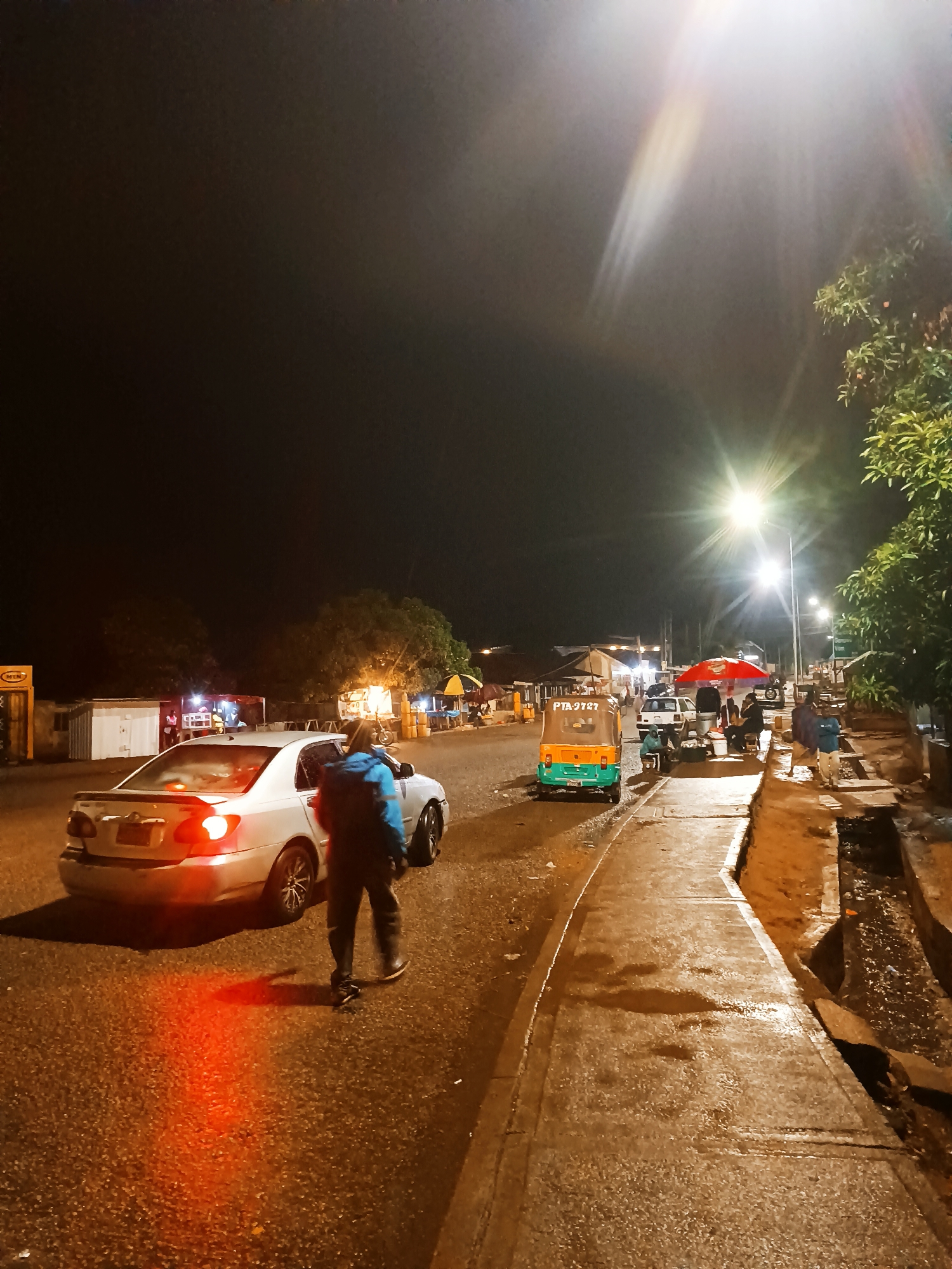 The image shows a street scene at night in Bukuru, Nigeria. A white sedan is on the left side of the street, with its brake lights illuminated. A man in a dark jacket with a blue backpack walks away from the car, toward the sidewalk. On the right side of the street, there's a green and yellow tuk-tuk with "PTA 9787" on it. The street is lined with streetlights. Several small businesses and people are visible in the background. The sidewalk is wet, and there's a visible drainage ditch.