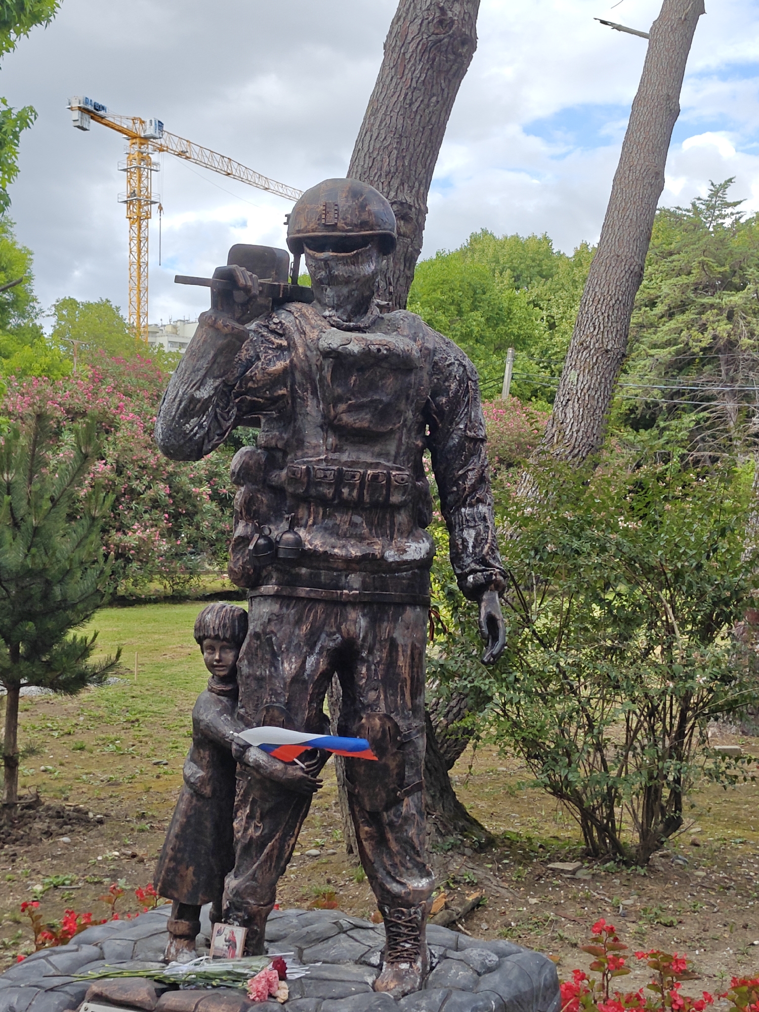 The image shows a bronze sculpture of a soldier carrying a rifle and a child holding a Russian flag. The sculpture is located outdoors in a park-like setting with trees and flowers. In the background, a yellow construction crane is visible. The sculpture appears to be a memorial, with flowers and a small framed image at its base. The soldier is depicted in military attire, including a helmet and body armor, while the child is shown in simpler clothing. The child is embracing the soldier's leg, and the flag is held between them.