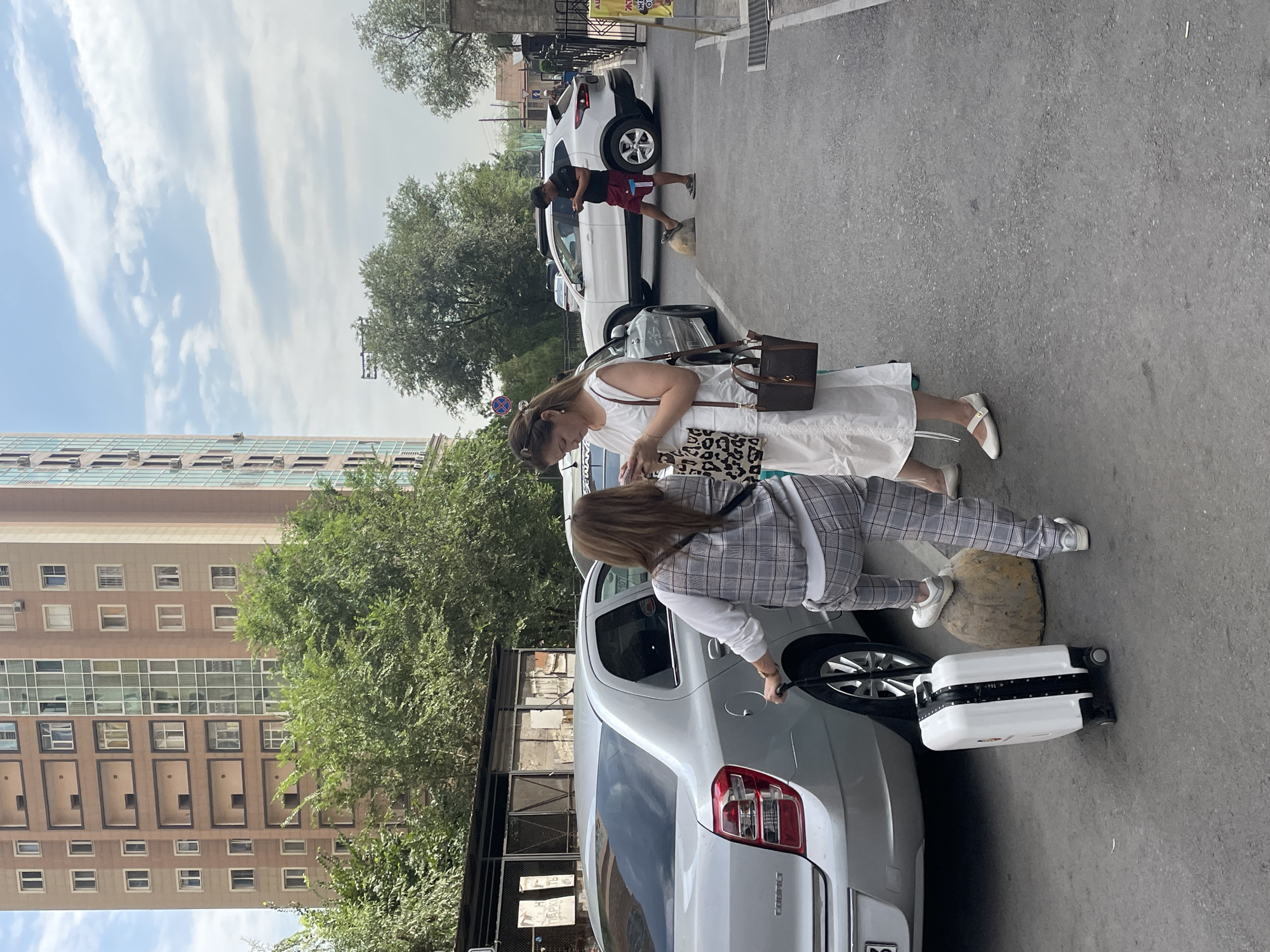 Two women and a man are present in the scene. The woman on the left is wearing a plaid vest over a white long-sleeved shirt and plaid pants, with white sneakers. She is holding onto the handle of a white rolling suitcase and has one foot on a large rock. The woman on the right wears a white sleeveless dress and white mary jane shoes. She is holding a brown handbag with a leopard print strap and appears to be looking at her phone. The man, positioned further back, is wearing red shorts and a dark t-shirt, with his phone in his hand and one foot on a similar rock.
They are standing on an asphalt surface next to a silver sedan. In the background, a modern, tall building with a beige and light brown facade and numerous windows is visible, along with green trees. A white SUV is parked behind the silver sedan. The sky is partially cloudy. The scene appears to be in Almaty, Kazakhstan, as indicated by the presence of a building that resembles architecture found in Almaty.