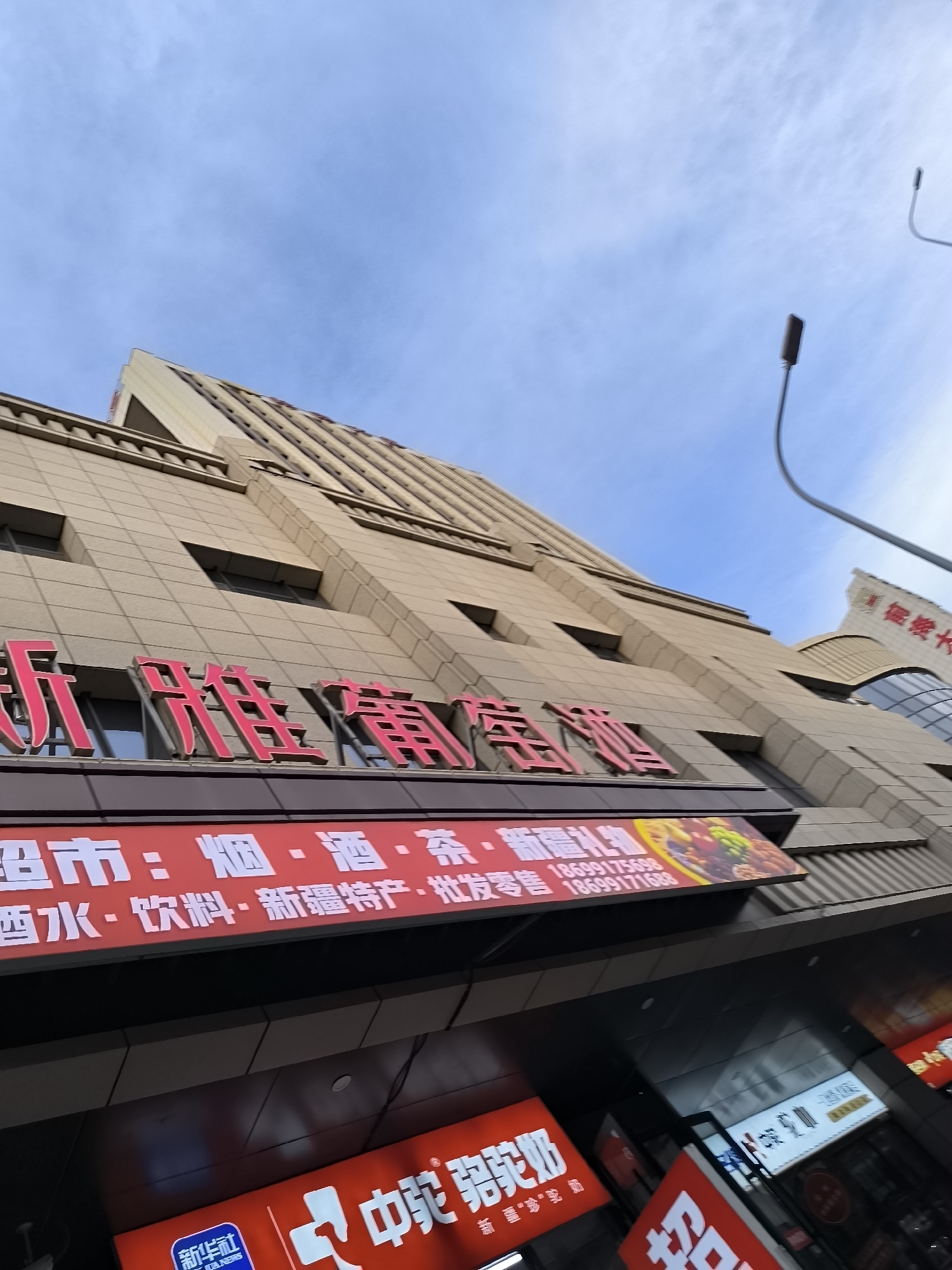 This is a ground-level exterior shot of a modern, multi-story building with a beige tiled facade, captured from a low angle looking upwards. The building has a distinctive stepped architectural design. Dominating the foreground and mid-ground are large, vibrant red Chinese characters on signs.
The primary red sign displays "新华雅苑商贸" (Xīnhuá Yǎyuàn Shāngmào), which translates to "Xinhua Garden Business and Trade." Below this, another red sign reads "超市:烟·酒·茶·香烟·酒水·饮料·新疆特产·批发零售" (Chāoshì: yān·jiǔ·chá·xiāngyān·jiǔshuǐ·yǐnliào·xīnjiāng tèchǎn·pīfā língshòu), meaning "Supermarket: Tobacco, Alcohol, Tea, Cigarettes, Alcoholic Beverages, Drinks, Xinjiang Specialties, Wholesale and Retail." This sign also features an image of assorted fruits and nuts, suggesting a diverse product offering. Two phone numbers are visible on this sign: 18699175698 and 18699171688.
Further down, two more red illuminated signs are partially visible. One displays white Chinese characters, the prominent ones being "中驼驼奶" (Zhōngtuó Tuónǎi), likely indicating camel milk products. The other sign has large white characters that appear to be "招" (Zhāo), meaning "recruit" or "invite."
The sky above is a clear blue with some wispy white clouds. A modern street lamp with a curved arm and a dark lamp head is visible on the right side of the frame, extending upwards into the sky. Another, more distant lamp post is also visible.
The building's architecture and the signage suggest a commercial establishment, likely a supermarket or a retail complex specializing in a variety of goods, including local Xinjiang products. The presence of "Xinjiang Specialties" on the sign firmly places this scene within Ürümqi, the capital city of the Xinjiang Uyghur Autonomous Region of China.