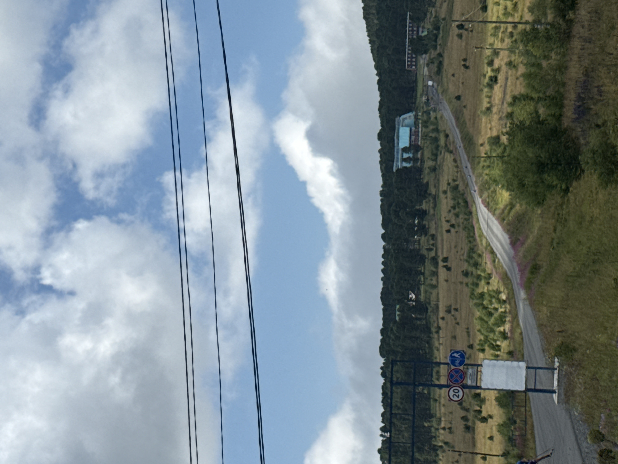 The image depicts a rural landscape under a cloudy sky. In the foreground, a road winds through rolling hills covered in dry grass and scattered bushes. A figure on skis is visible on the left side of the road, near a blue metal signpost with traffic signs and a blank white panel. The road leads towards a distant forested hillside. On the hillside, two buildings are visible: a larger, light-blue structure with a distinctive curved roof, and a smaller, darker rectangular building. A series of power lines stretches across the upper portion of the image. The overall impression is one of a remote, possibly mountainous, area.