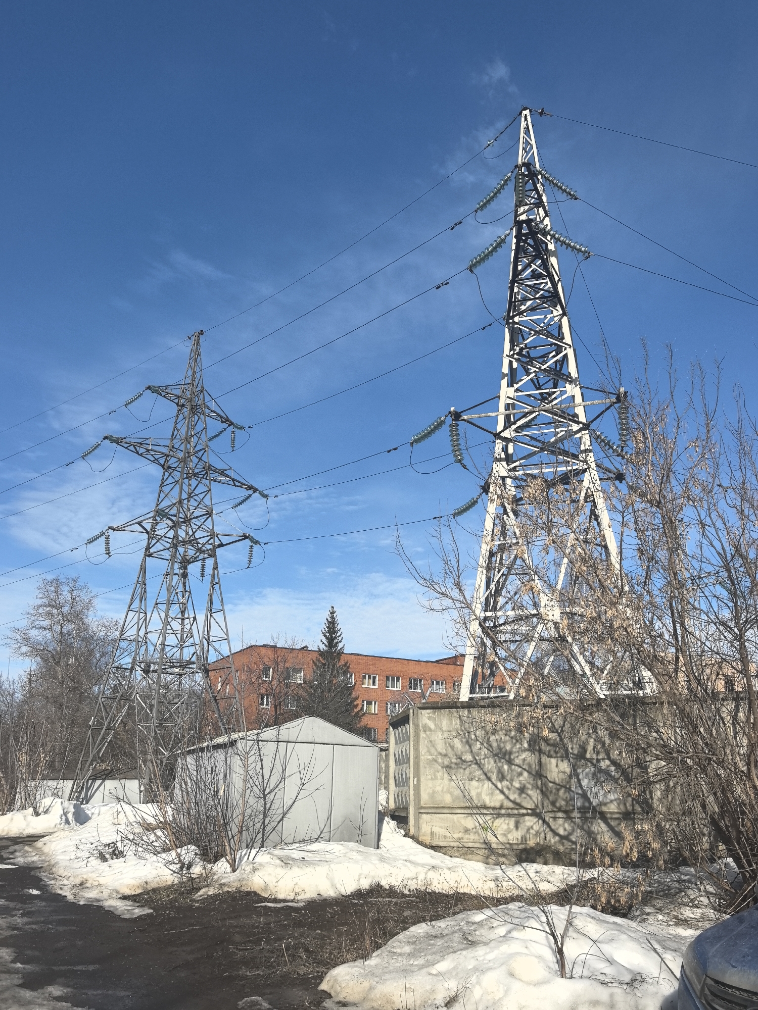 The image depicts an outdoor scene in Sarapul, Russia, characterized by two prominent lattice steel electrical transmission towers under a bright blue sky with minimal cirrus clouds. The ground in the foreground consists of dark, wet earth and substantial patches of melting snow, interspersed with numerous bare deciduous bushes and trees, some retaining dried leaves. A dark, possibly asphalt or dirt path, is visible in the lower-left. A portion of a light blue or silver vehicle is present in the lower-right corner.
The taller transmission tower, positioned to the right, is light gray or white and features multiple sets of light green/cyan disc insulators supporting numerous power lines that traverse the frame. A second, shorter, darker gray lattice tower stands to the left, also equipped with disc insulators and connected to power lines. In the mid-ground, a light gray, gabled-roof shed-like structure is visible near the base of the taller tower, and to its right, a concrete wall or utility structure with a patterned surface is present, casting tree shadows. In the background, a multi-story, reddish-brown brick building with white-framed windows is partially obscured by the bare trees and a solitary conifer tree. No people are visible.