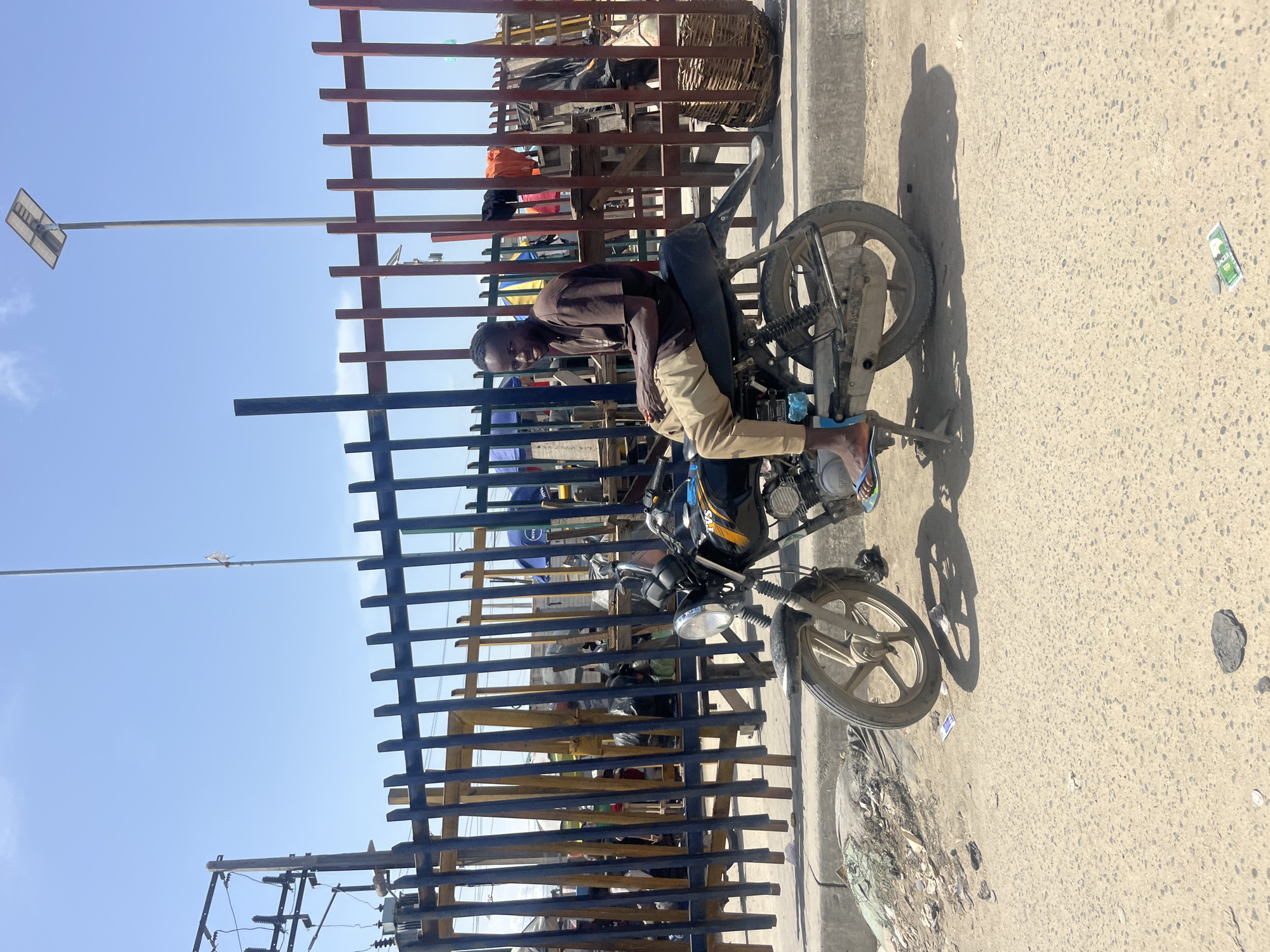 A man sits astride a black and gold motorcycle on a concrete surface next to a metal fence in Ado, Nigeria. The man is wearing a brown shirt, khaki pants, and blue flip-flops. He has a dark complexion and is looking towards the camera. The motorcycle is parked adjacent to the curb. Debris is scattered on the ground next to the curb. The fence is made of horizontal metal bars painted blue and brown. A bright blue sky is overhead. Overhead, electrical poles and power lines are present. A streetlight is visible in the background.