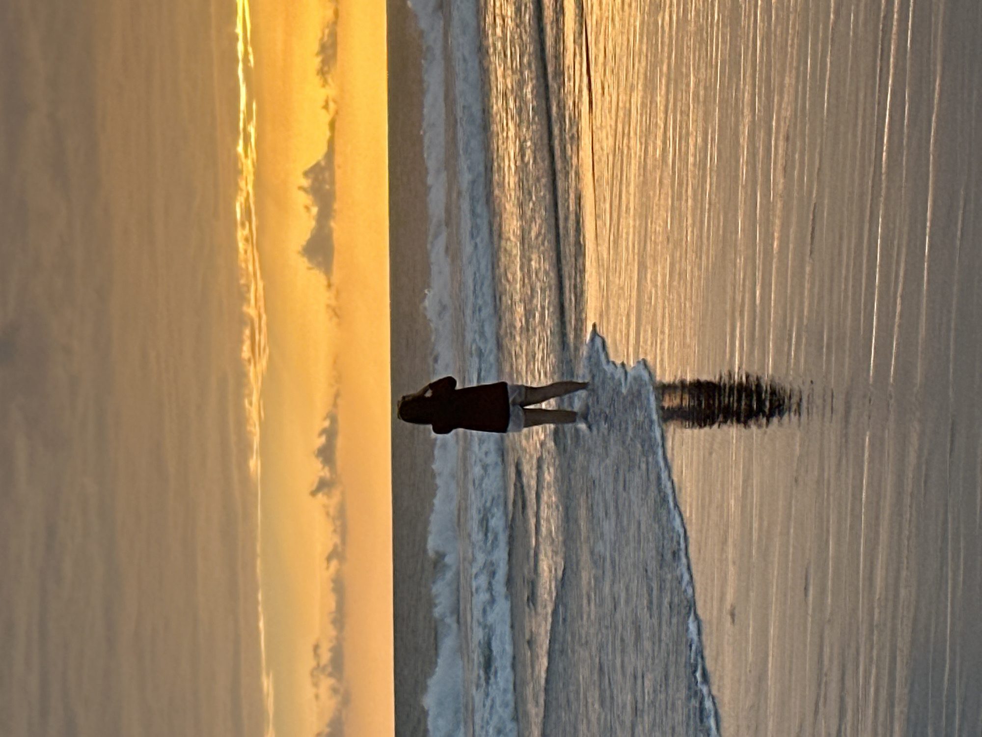 A lone figure stands in shallow water at the edge of the ocean in North Kuta, Indonesia, facing the horizon. The sun is low in the sky, casting a warm, orange glow on the scene. The person, wearing a dark top and light-colored shorts, appears to be taking a photo with a phone. Waves are breaking in the background, and the wet sand reflects the light from the sunset. The sky is filled with clouds, some illuminated by the setting sun.